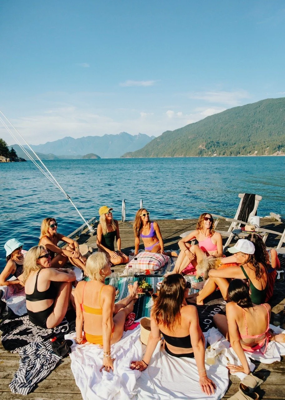 Group of women enjoying a sunny day on a wooden dock by the water, sitting on blankets and cushions, surrounded by mountains and a lake.