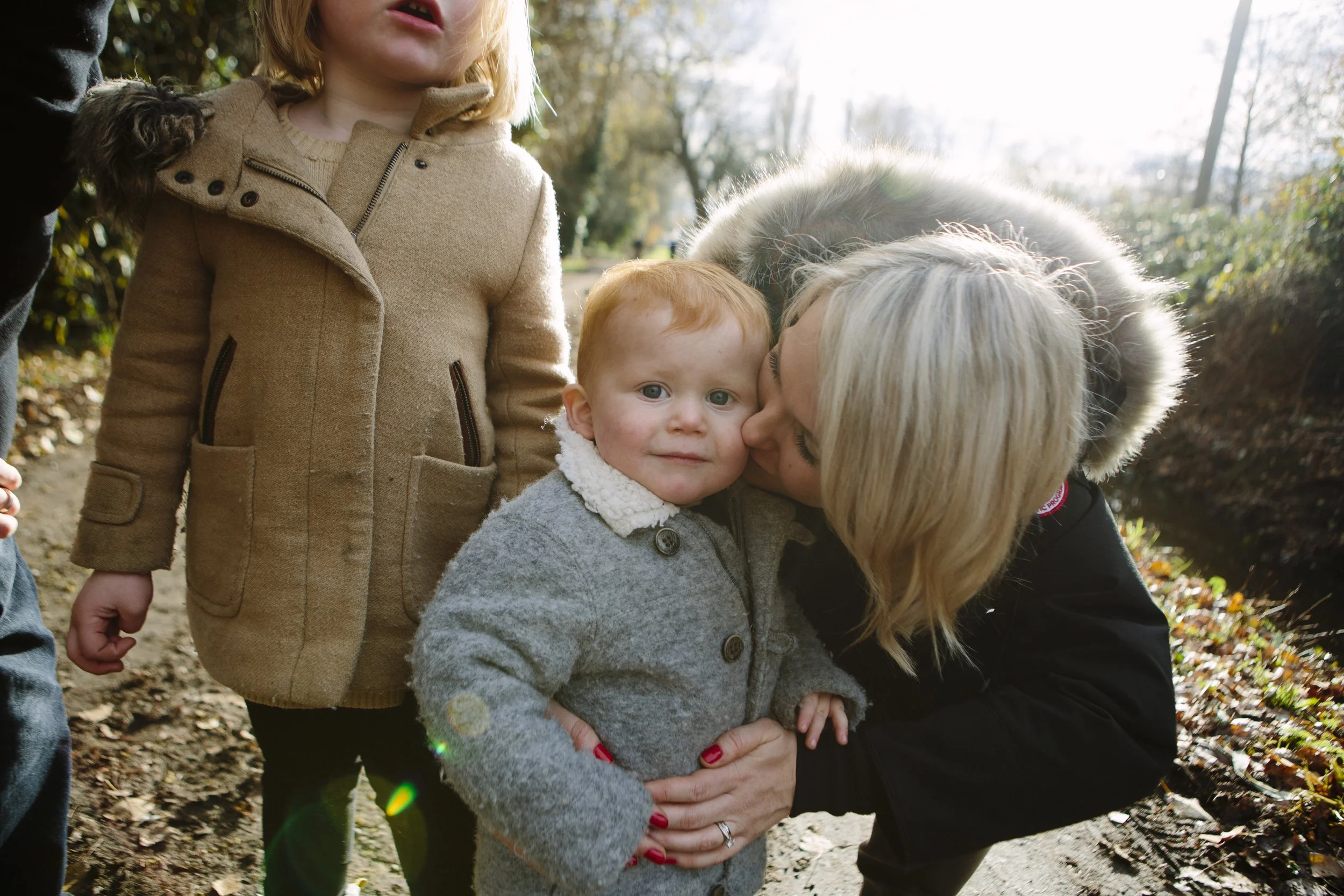 A woman is kissing a young boy on the cheek while holding him. There is also a girl in a tan coat and a person on the left side of the image. They are outside on a trail surrounded by trees.