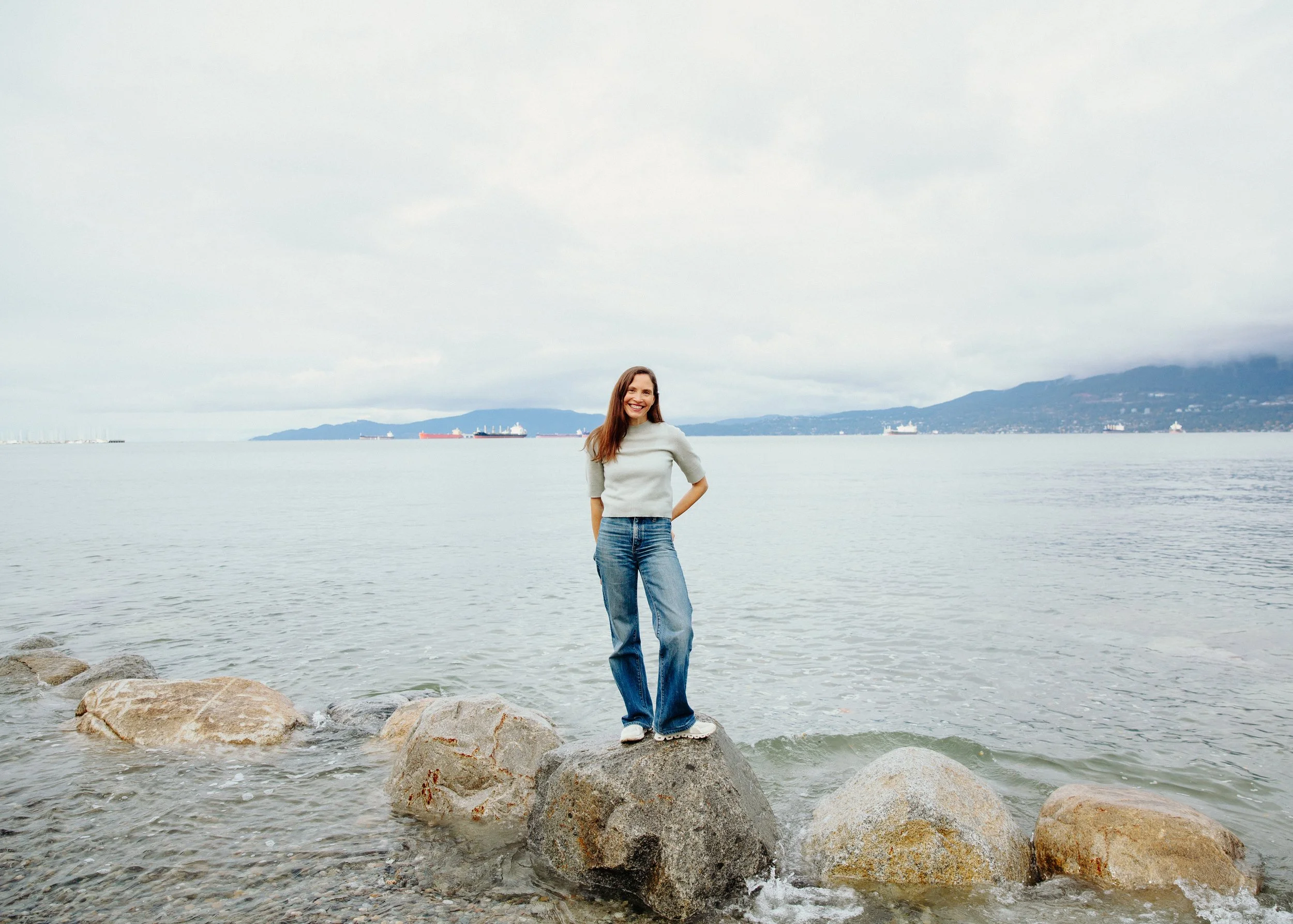 A woman standing on rocks by the water with a scenic background of hills, ships, and a cloudy sky.