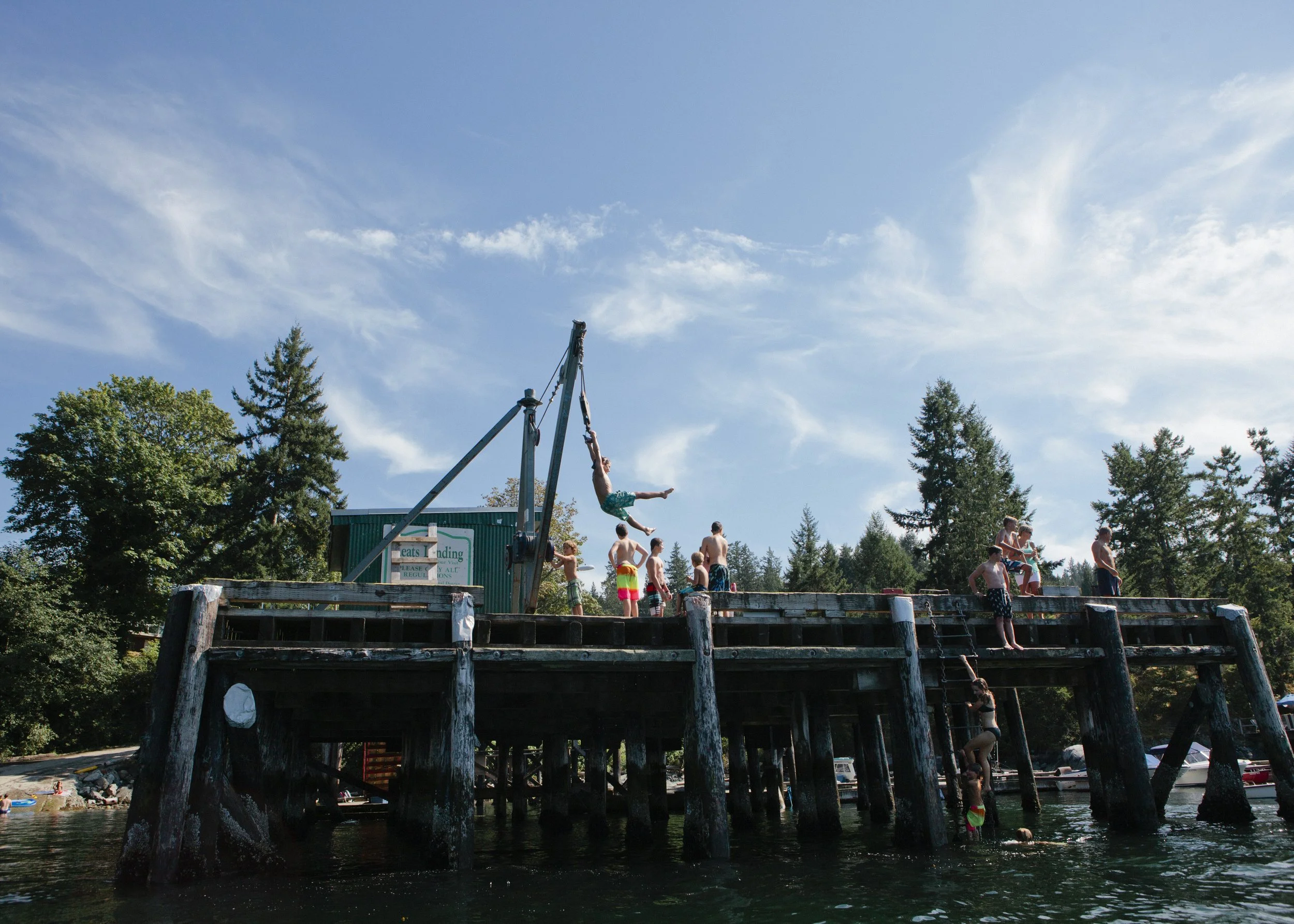 People enjoying a day at a water park with a large wooden pier and a diving board, green trees in the background, and a clear blue sky with clouds.