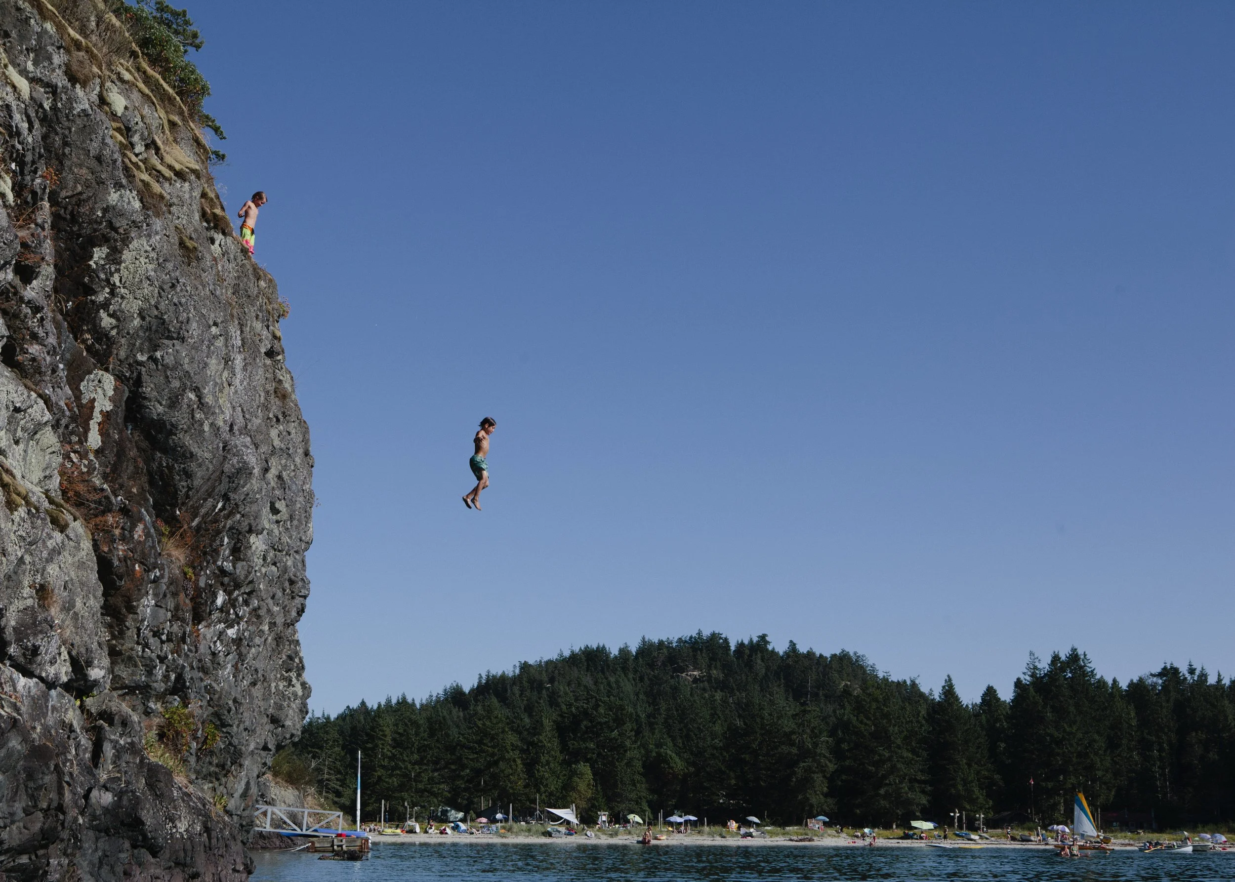People jumping off a cliff into a lake surrounded by forest