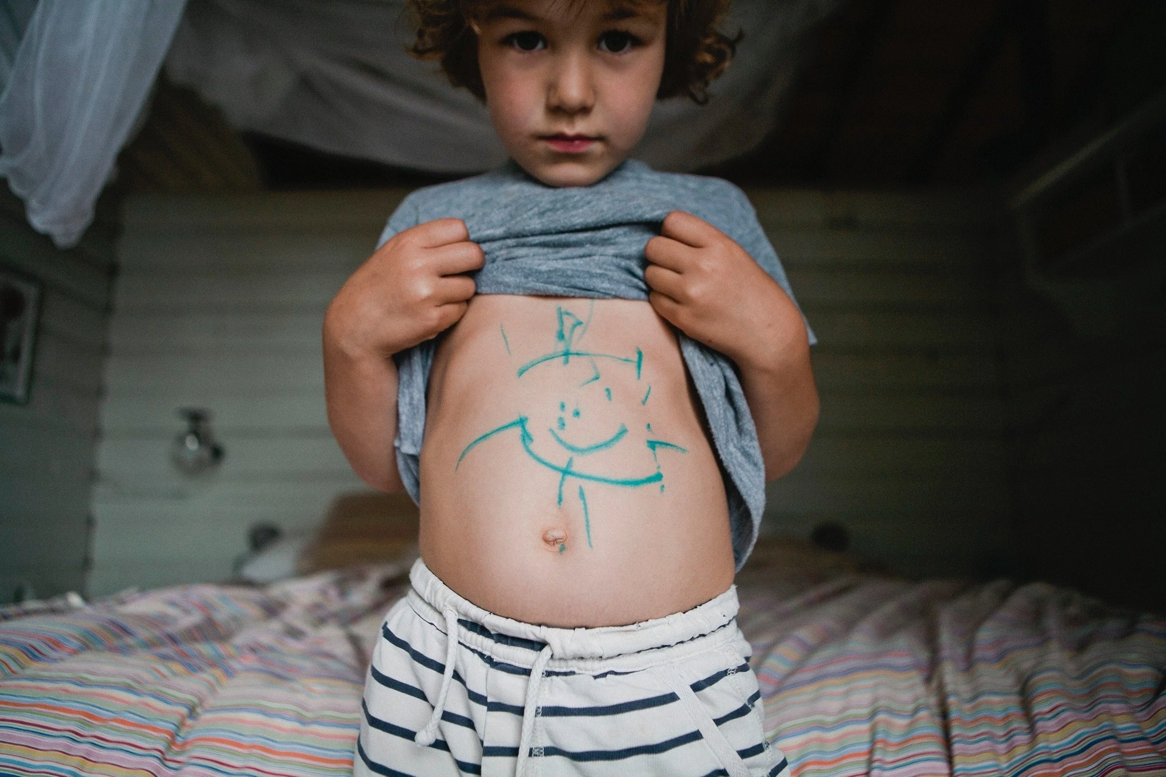 A young boy with curly hair lifts his gray shirt to reveal his abdomen, which has blue marker drawings of a sun, clouds, and other shapes. He stands in front of a bed with a colorful striped blanket.