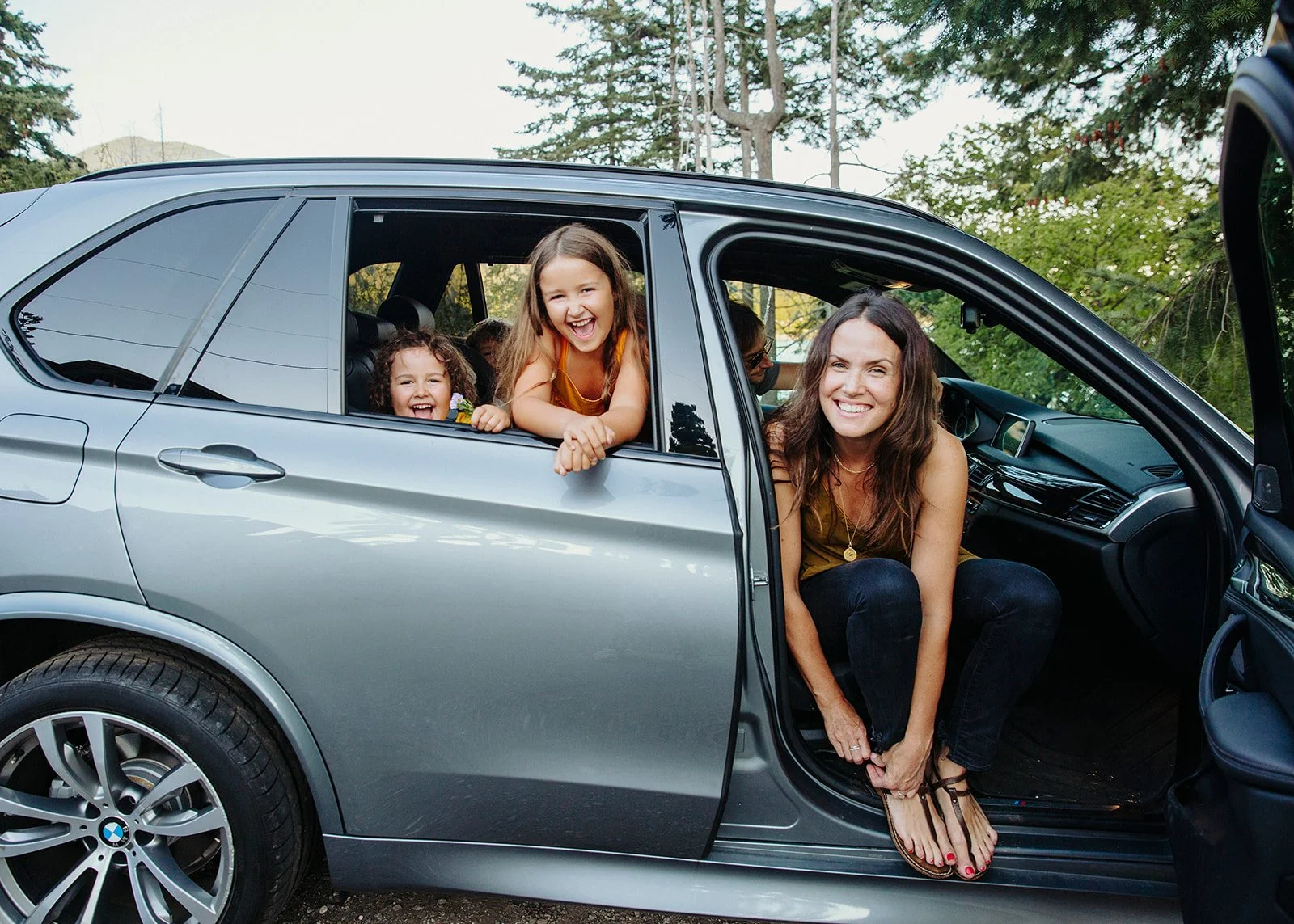 A family of four smiling in a parked gray BMW SUV, with two young girls leaning out of the open back window and their mother sitting on the edge of the front passenger seat.