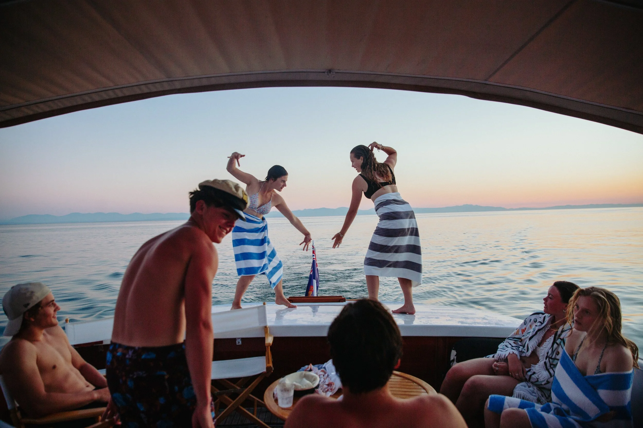 People dancing on a boat at sunset, with water and distant mountains in the background.