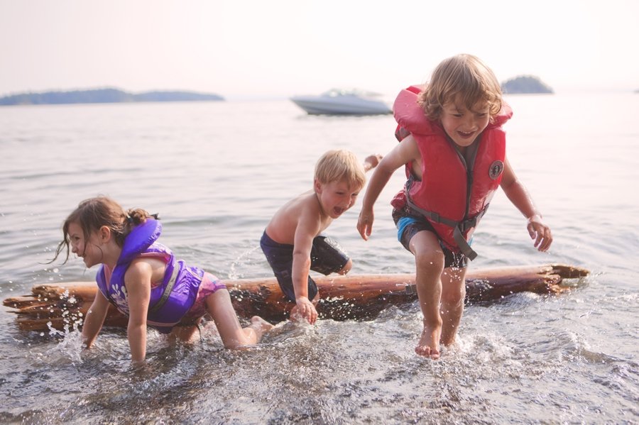 Three kids playing on a surfboard in water near a boat, wearing life jackets.