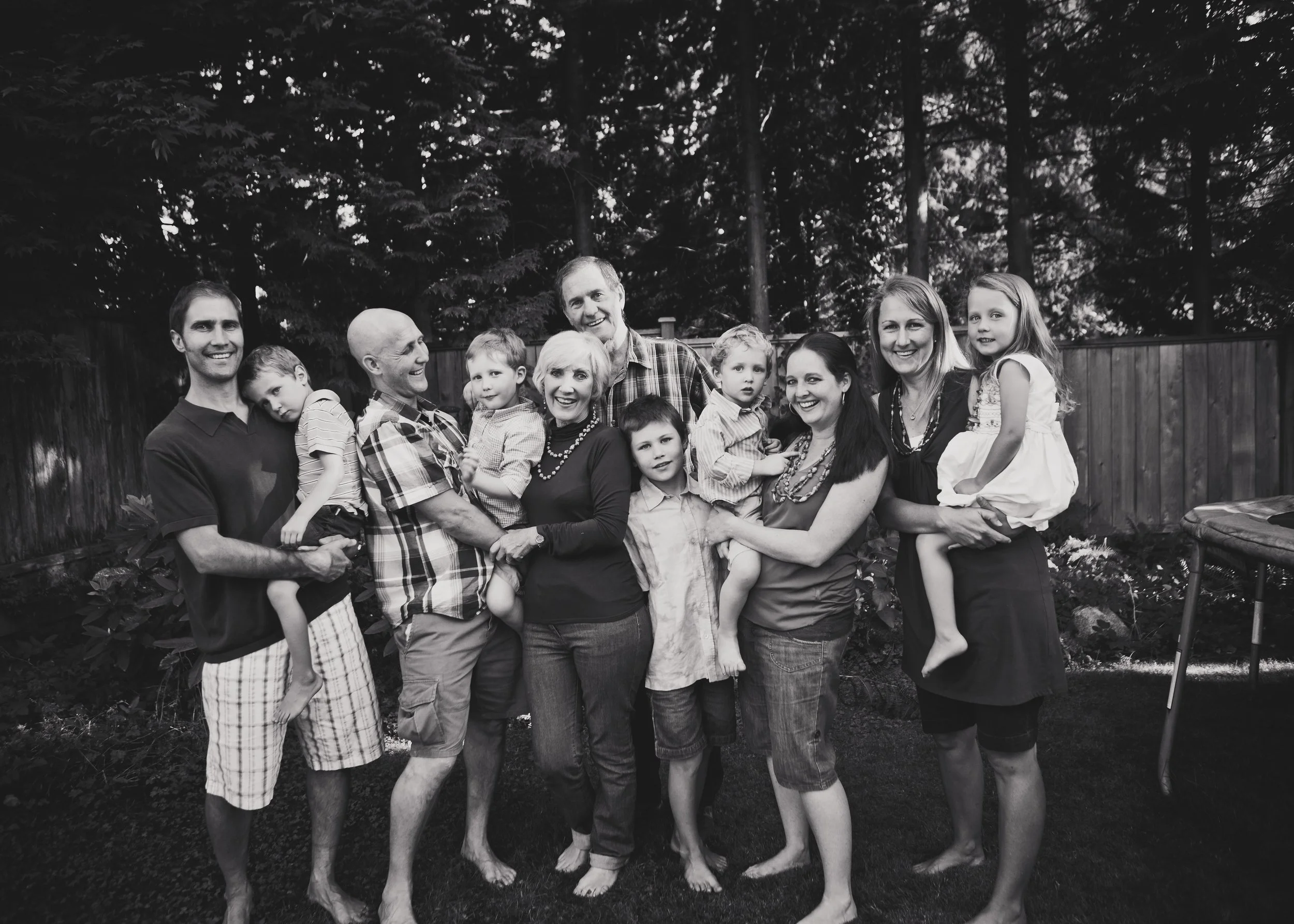 A black-and-white photo of a large family standing outdoors in a backyard, smiling and hugging each other.