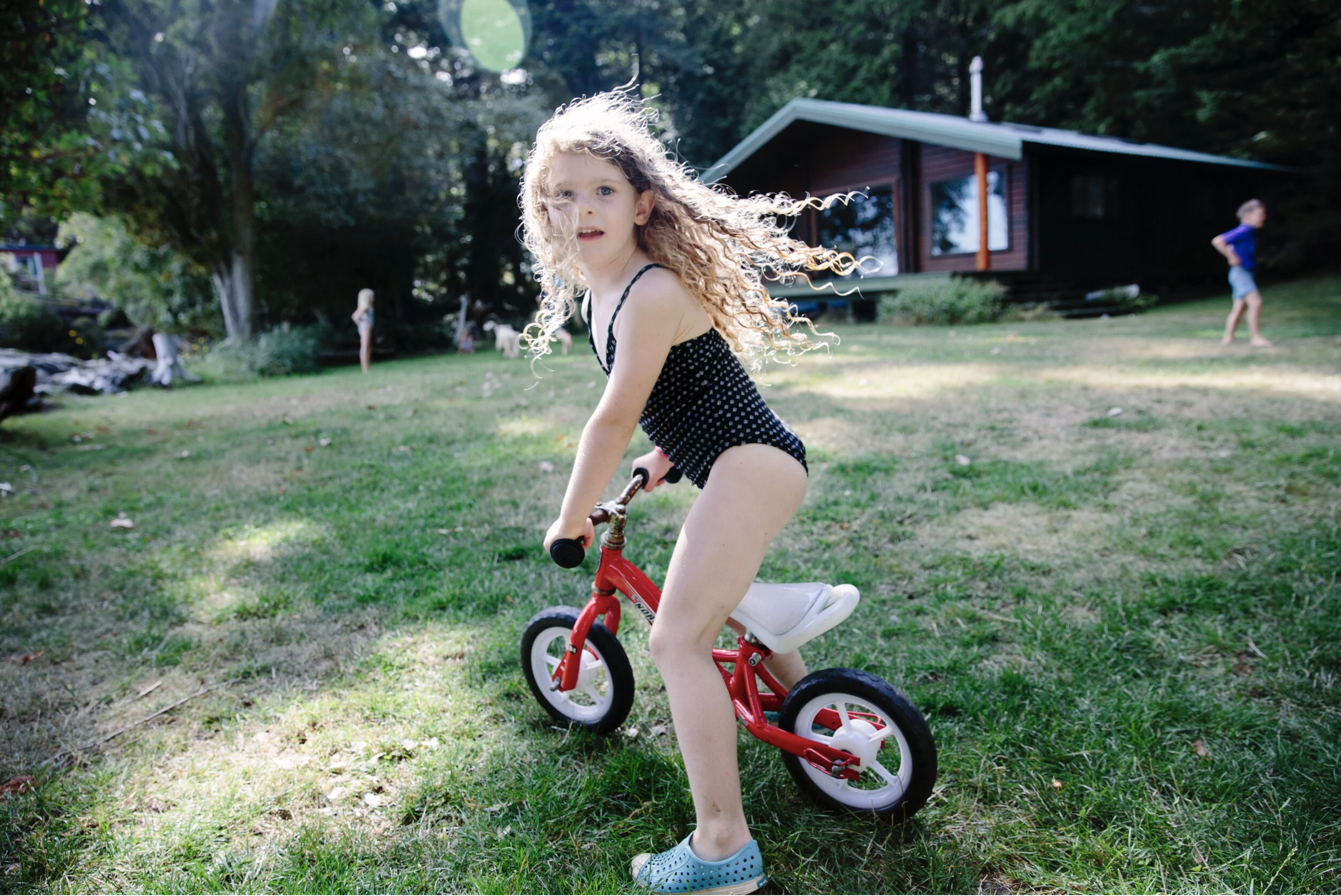 A young girl with curly blonde hair riding a small red and white bike outdoors on grass, with a treehouse and other children in the background.