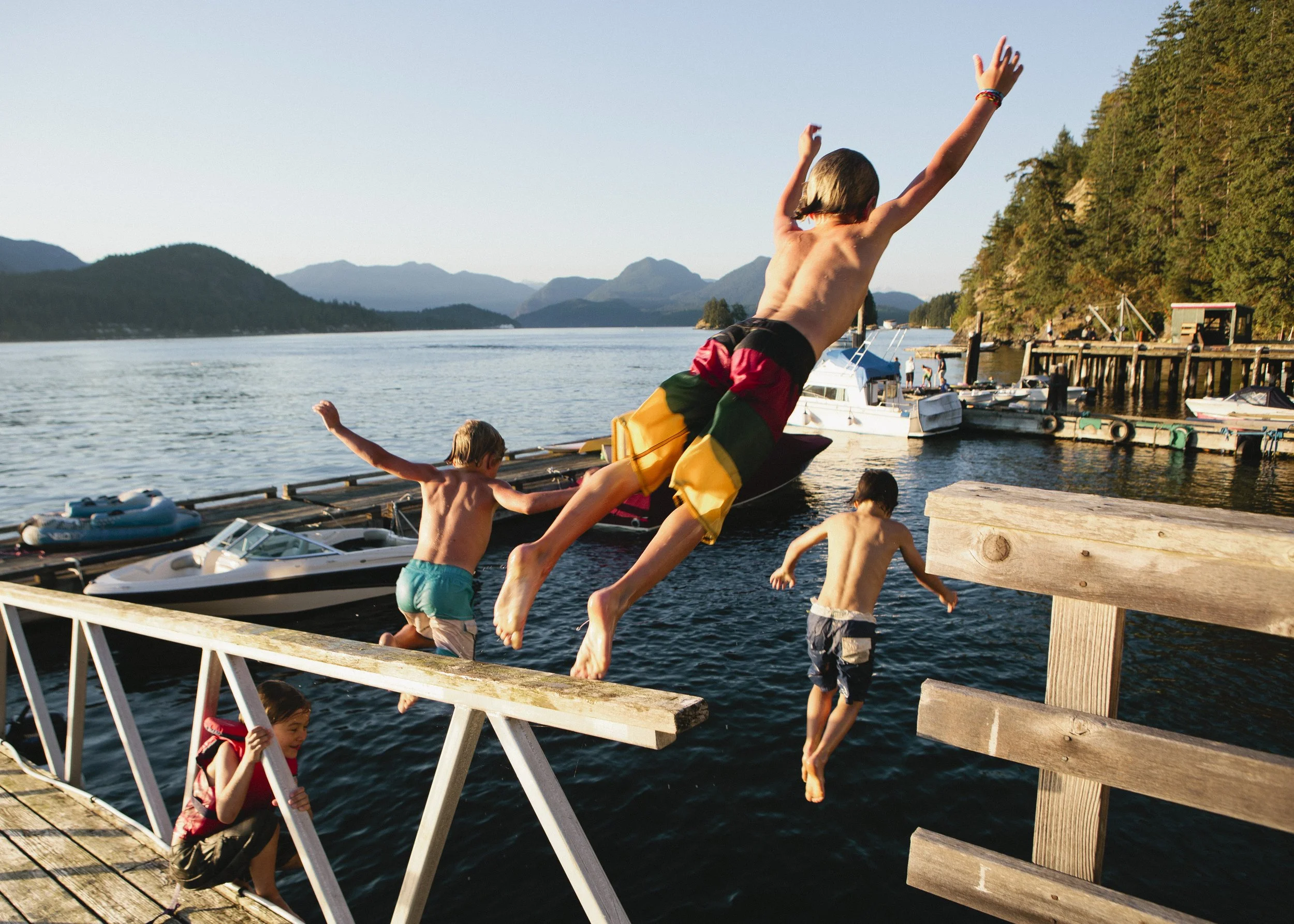 Children jumping off a wooden dock into a body of water, surrounded by boats, trees, and mountains in the background during late afternoon.