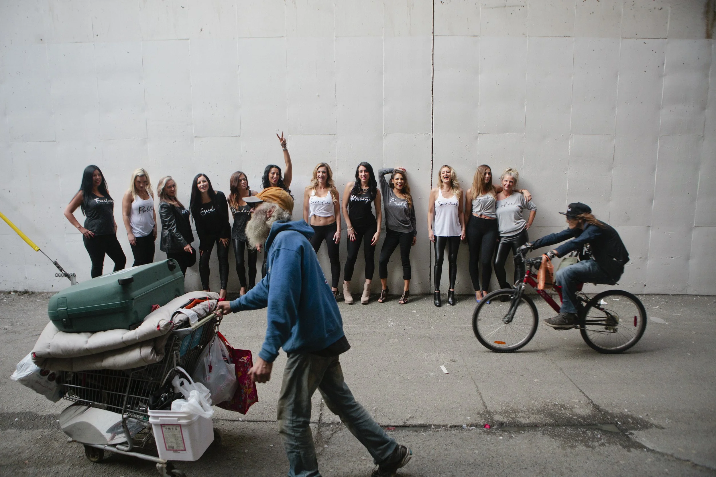 A group of nine women standing against a plain white wall, posing and smiling, with two people in the foreground and a person riding a bicycle on the right. The women are wearing casual tops with words like 'family', 'blessed', 'sacrifice', and 'spir