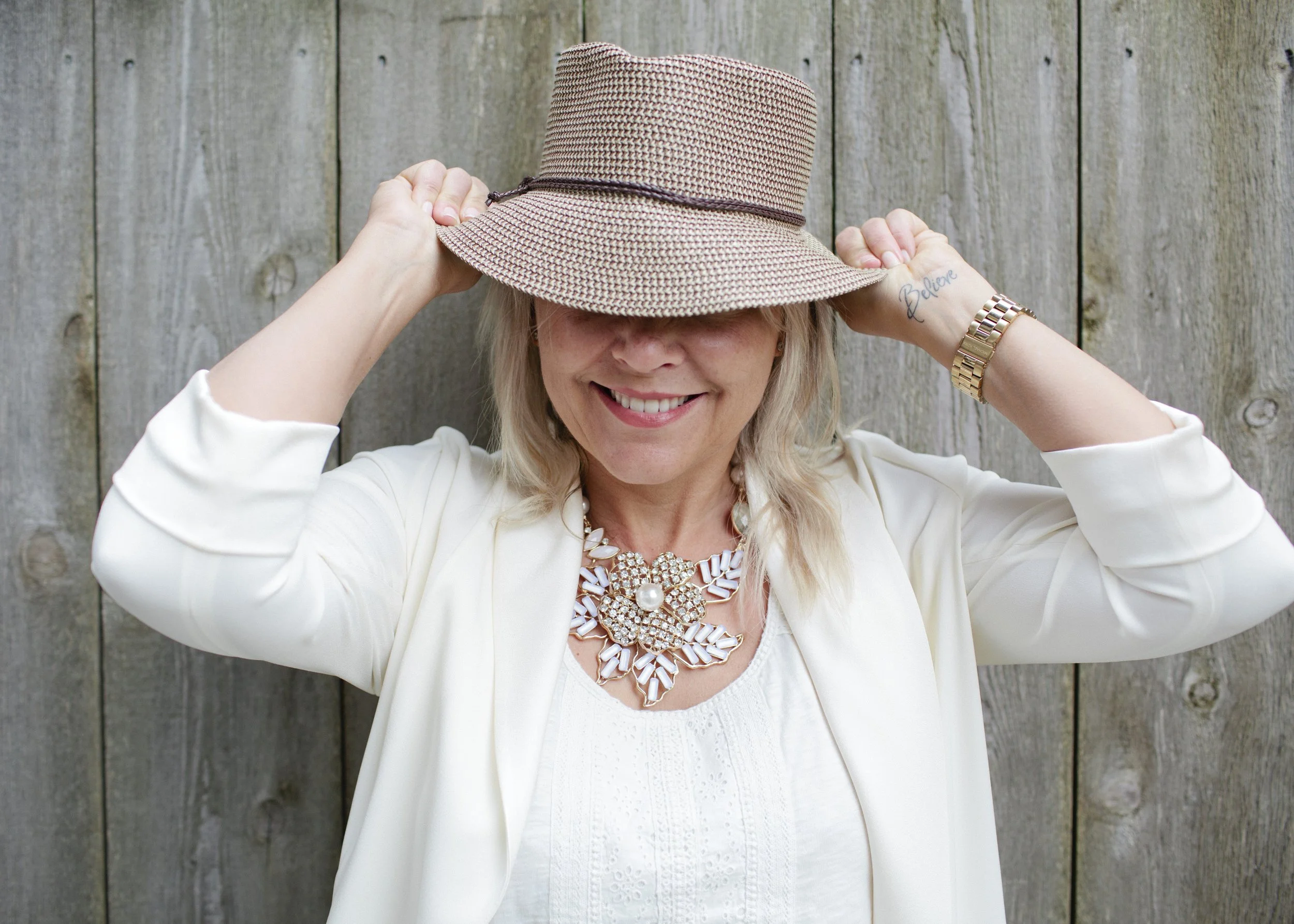 A smiling woman wearing a large beige sunhat, white blazer, and statement jewelry, standing outdoors in front of a wooden fence.