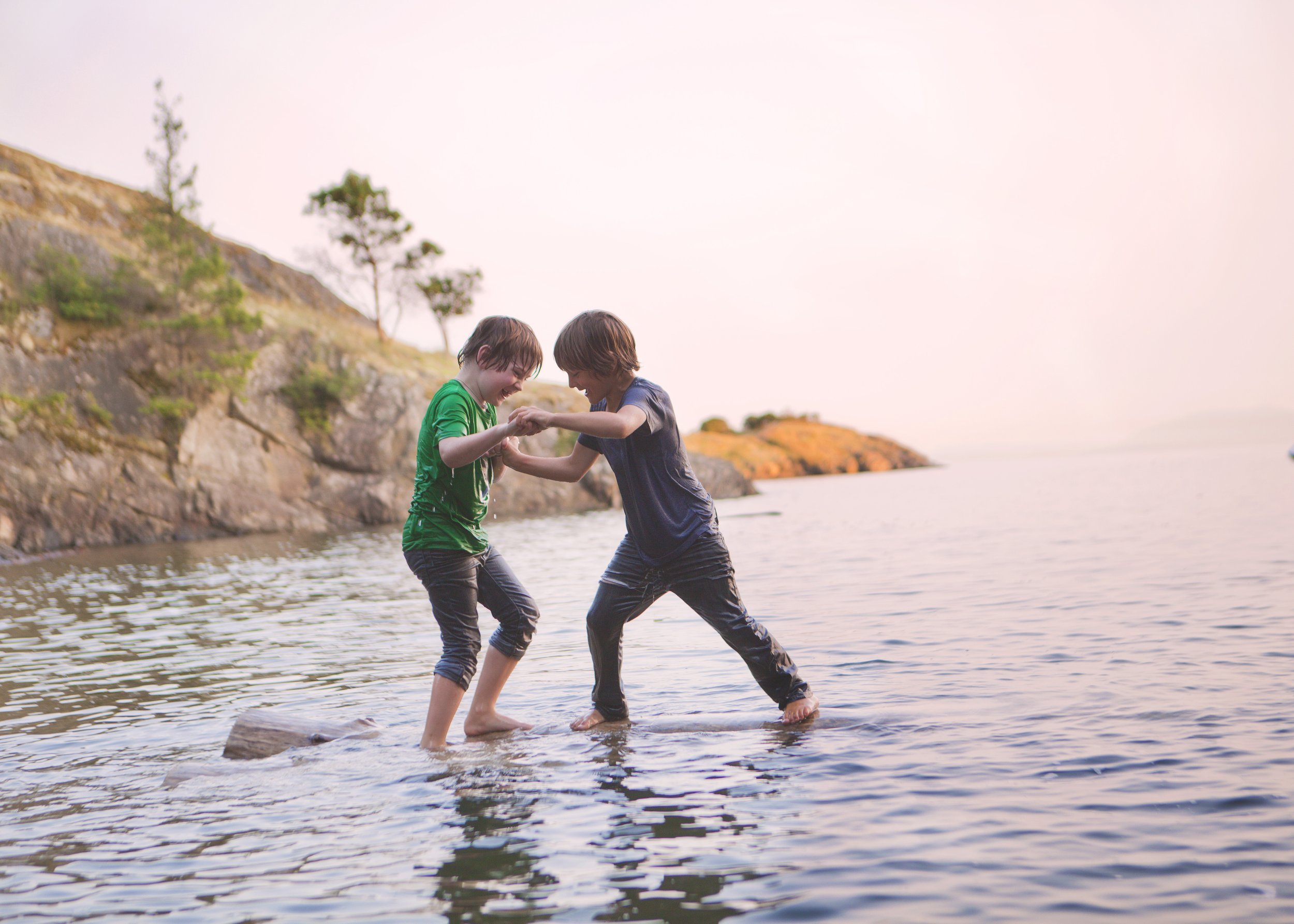 Two children are playing and splashing water on a rock in the ocean near a rocky shoreline with a few trees, during sunset or sunrise.