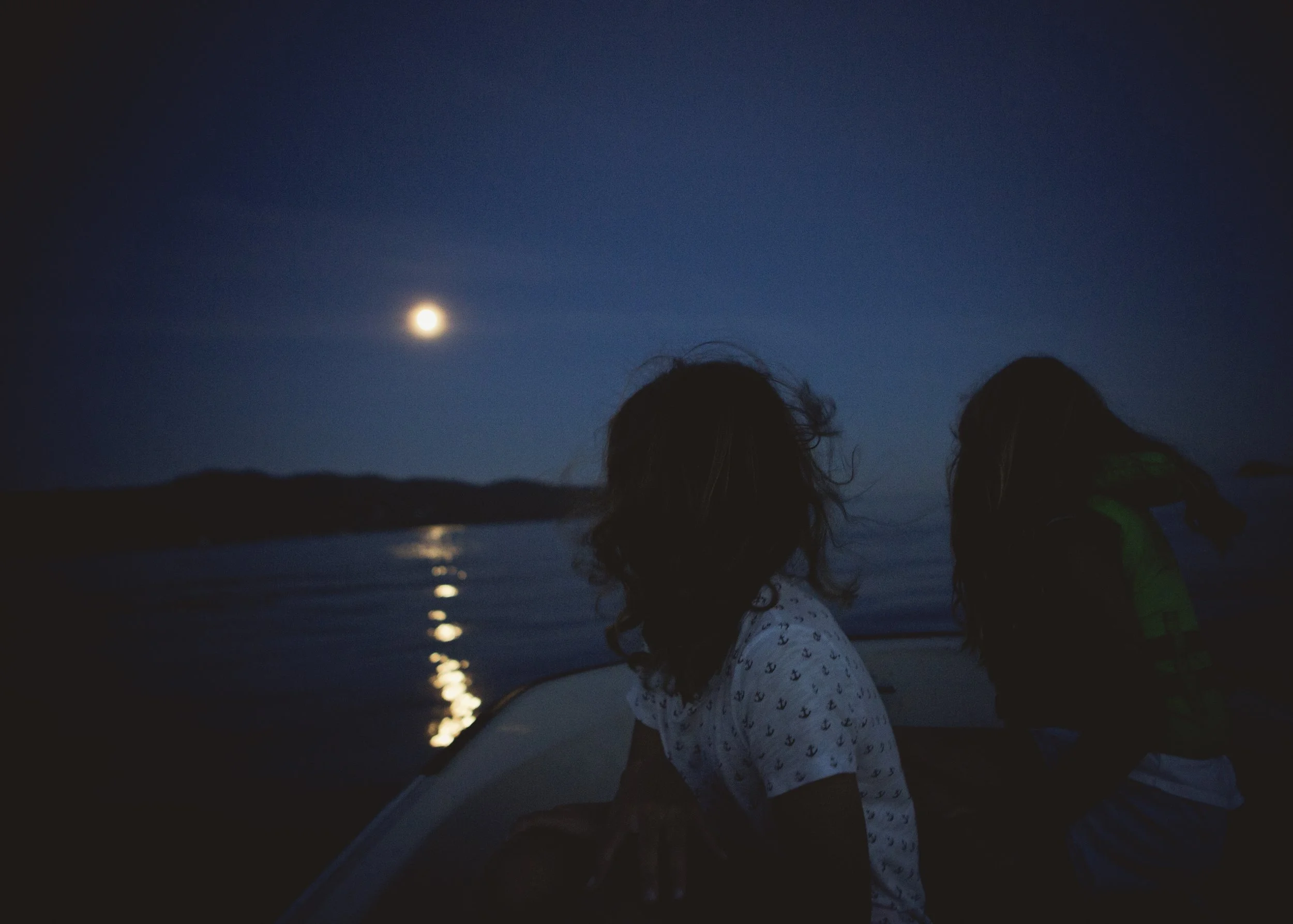 Two children stand by a body of water at night, illuminated by the moon in the darkening sky.