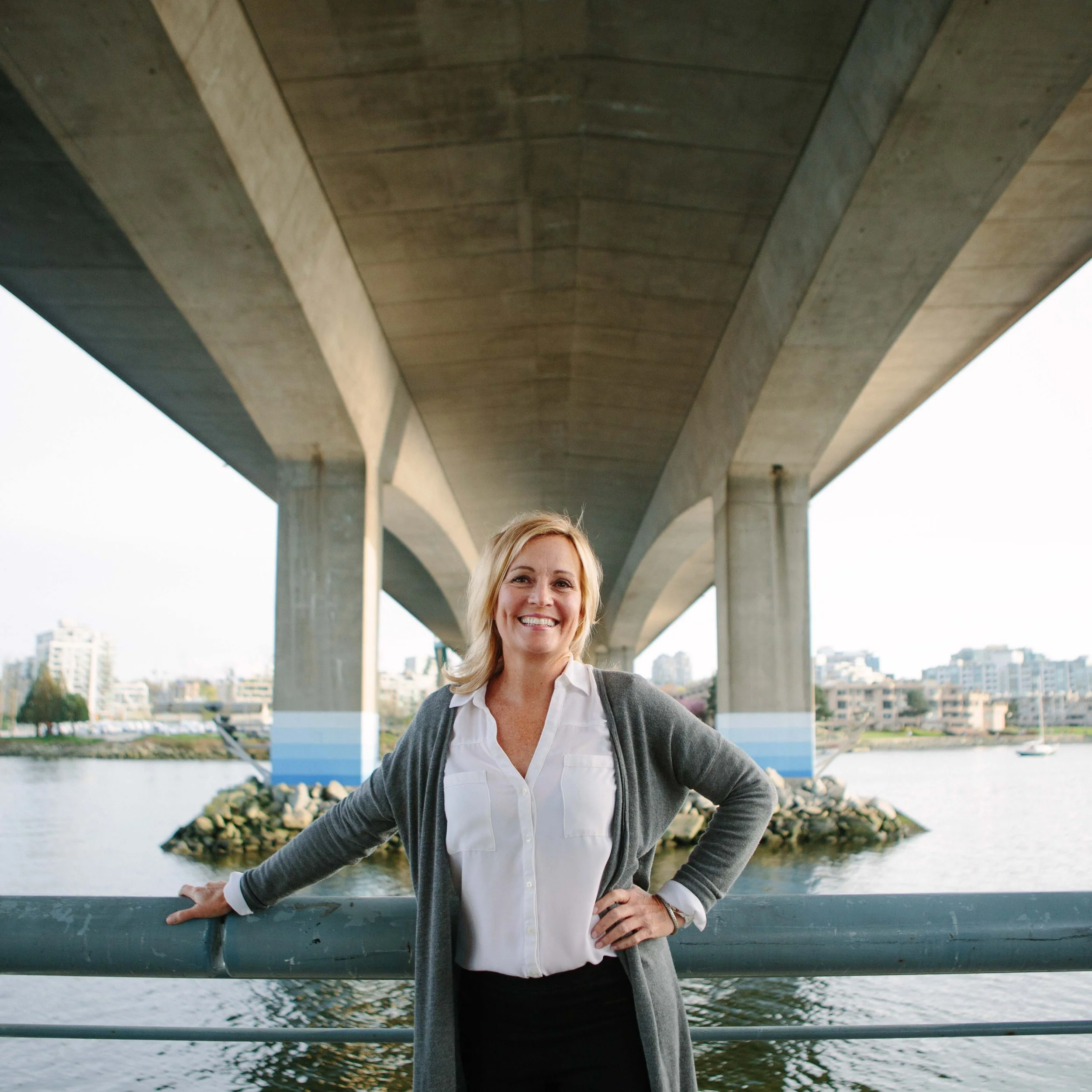 A woman with blonde hair smiling under a large concrete bridge by a river, wearing a white shirt, gray cardigan, and black pants.