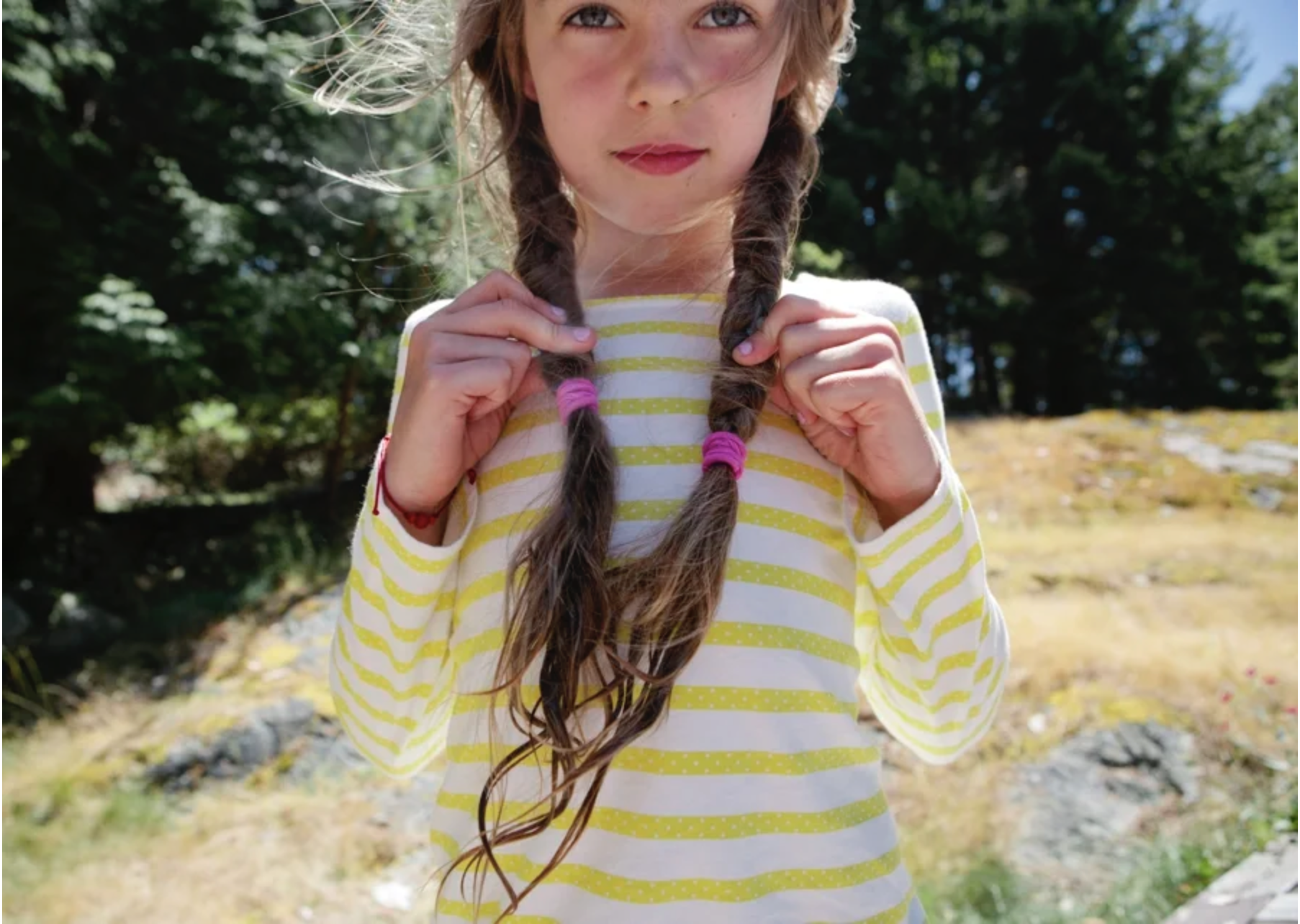 A young girl with tan hair in braids, wearing a yellow and white striped long-sleeve shirt, holding her braids with both hands, standing outdoors with trees and grass in the background.