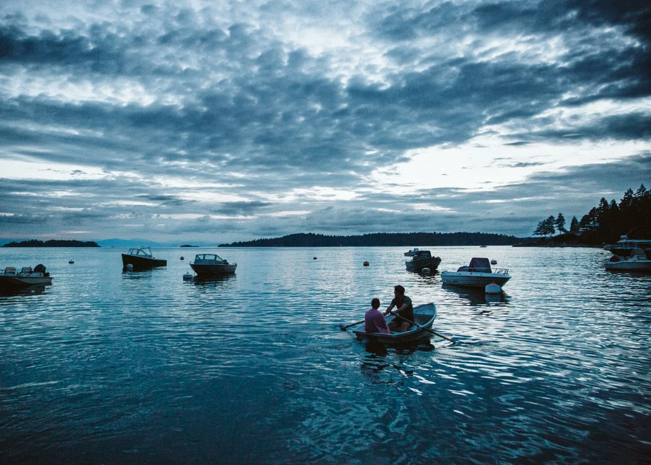 Two people boating on calm water with multiple boats anchored nearby under a cloudy sky.