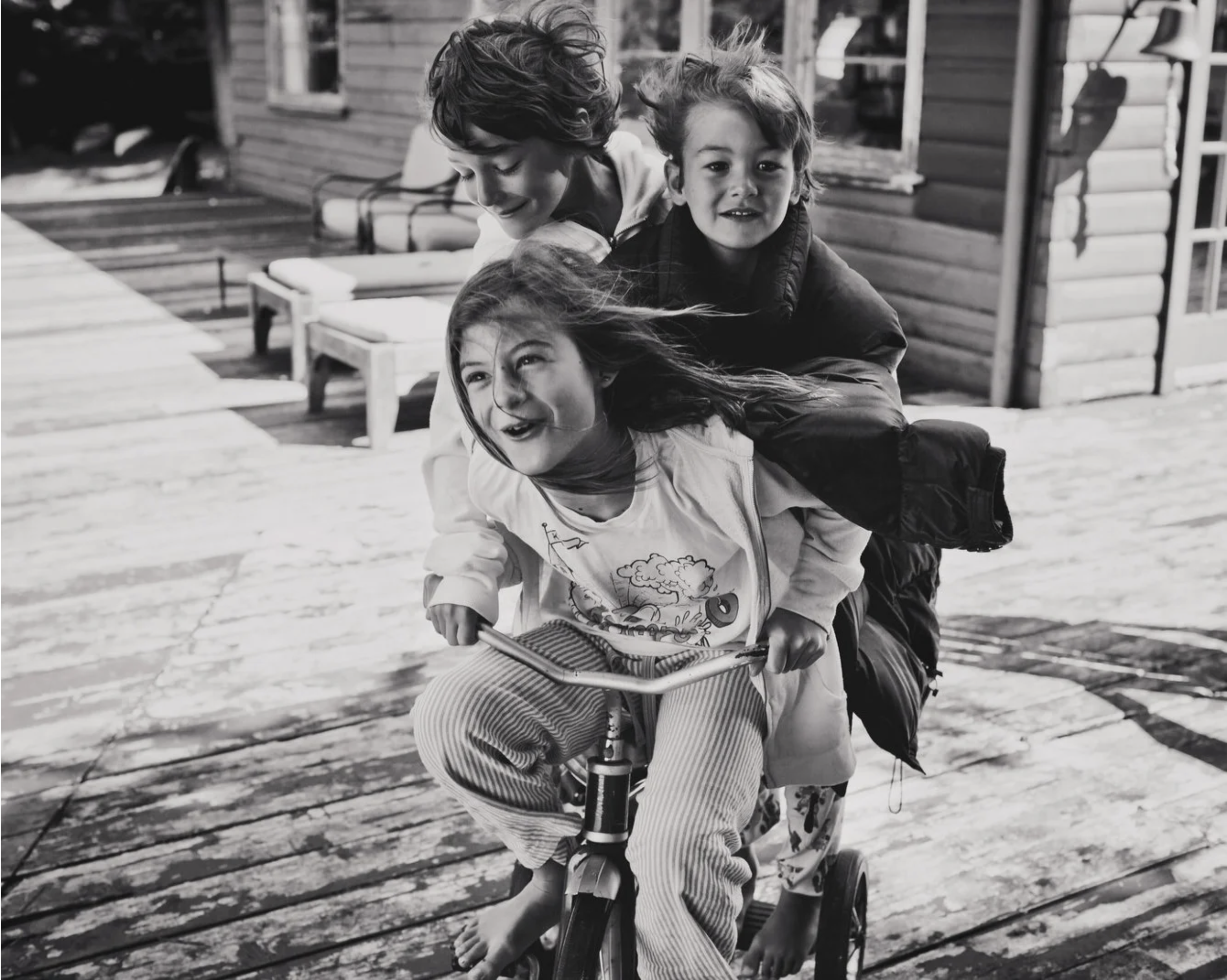 Three children riding a bicycle in a snowy outdoor area with a wooden building in the background, black and white photo.