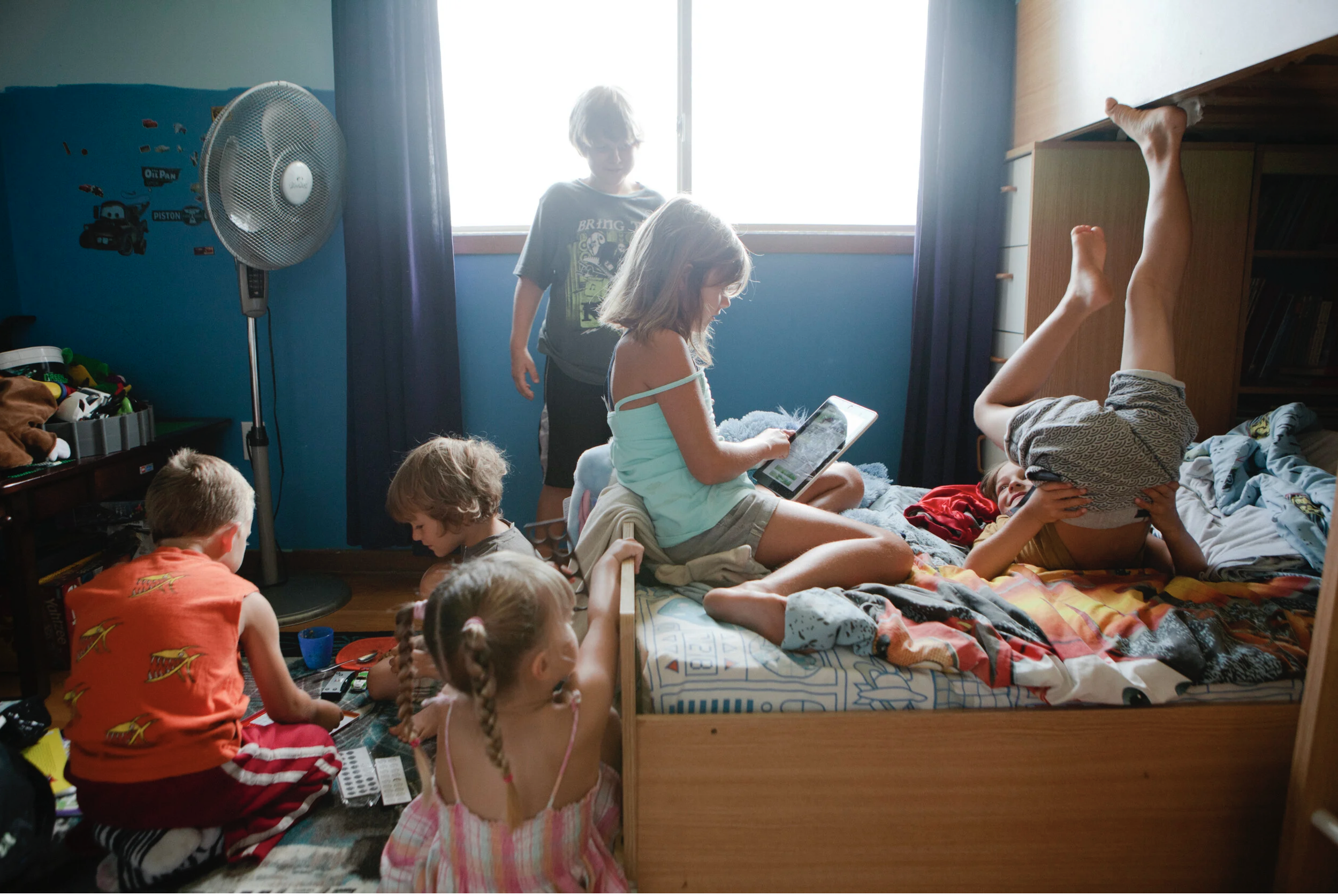 Several children are in a bedroom, some sitting on the floor playing with toys, while others are on a bed. One girl is sitting on the bed, using a tablet, and another girl is lying upside down on the bed. In the background, a boy is standing near a w