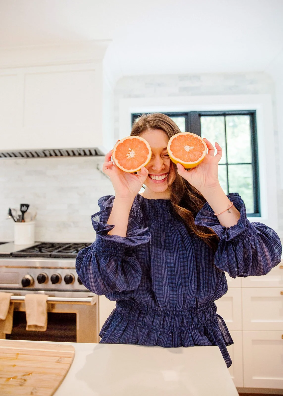 A woman smiling in a kitchen holding two halves of a grapefruit in front of her eyes.