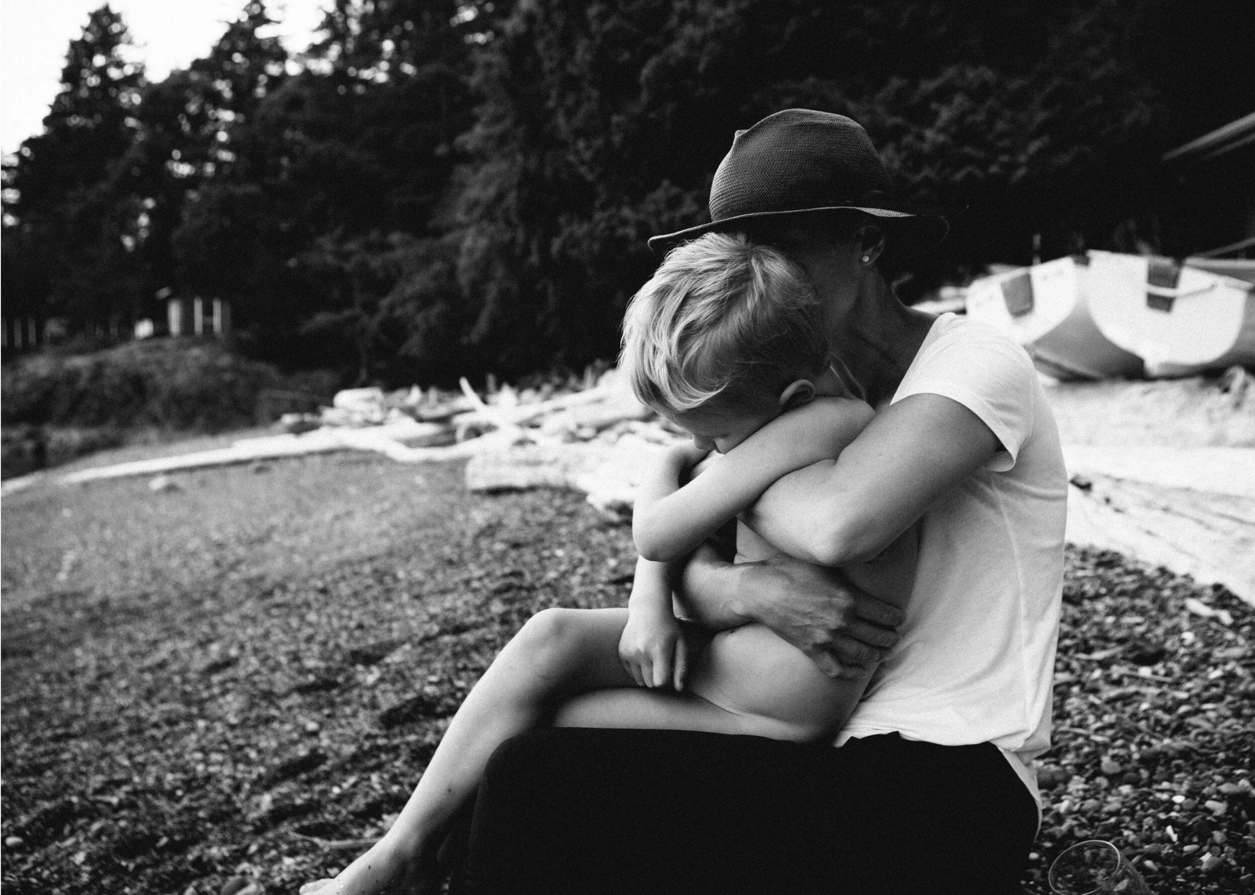 A person with short blonde hair hugging a woman wearing a hat outdoors by a body of water with trees in the background.