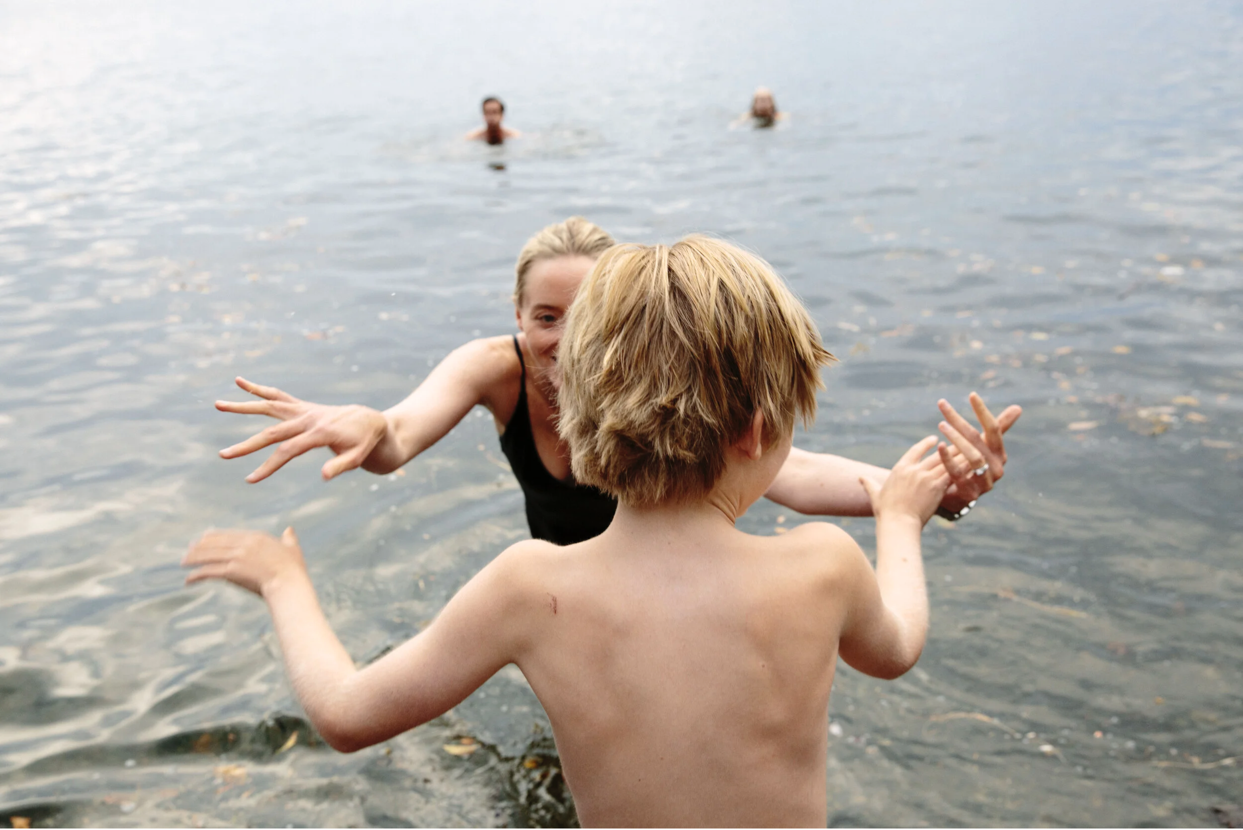 People swimming and playing in a body of water, seen from behind a child with blonde hair in the foreground, with a woman and another person in the background.