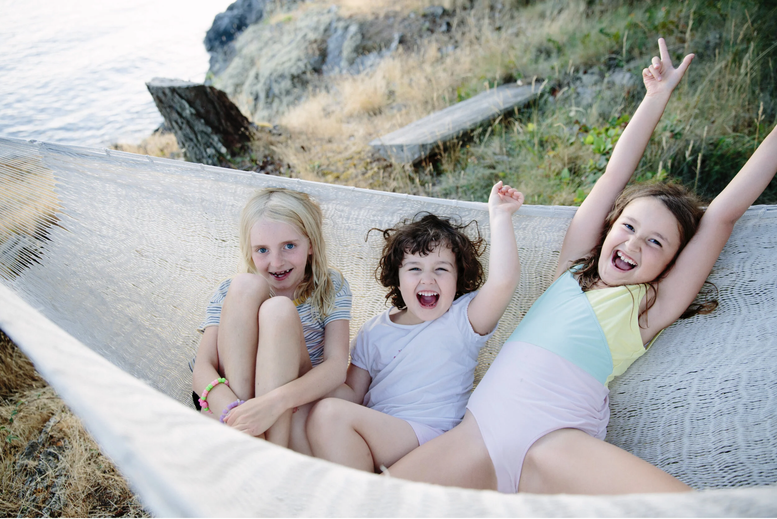 Three children relaxing in a hammock near a rocky coastline with water in the background. The children are smiling and making playful gestures.
