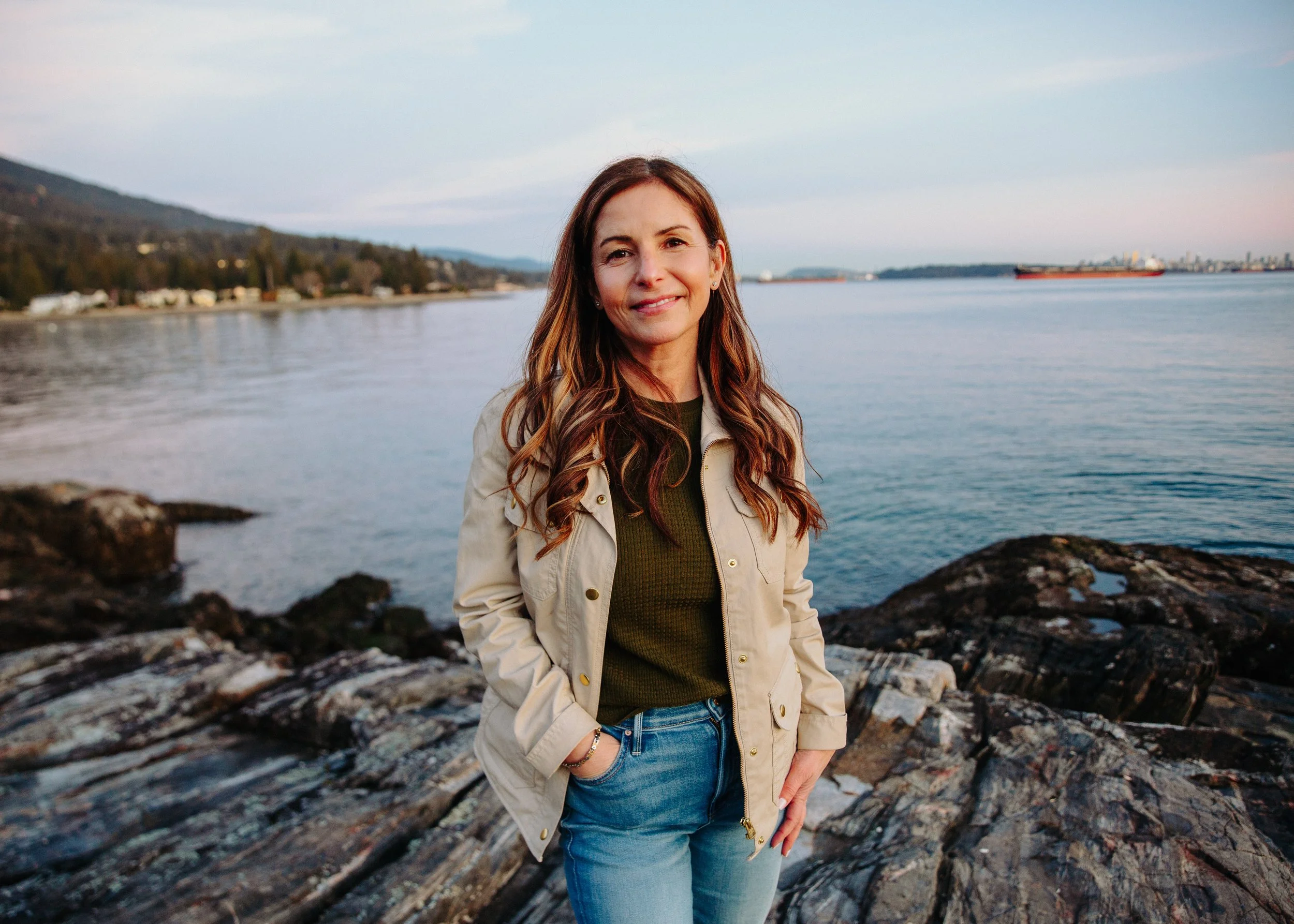 A woman with long wavy brown hair smiling at the camera, standing on rocks by the water with boats and hills in the background during the evening.