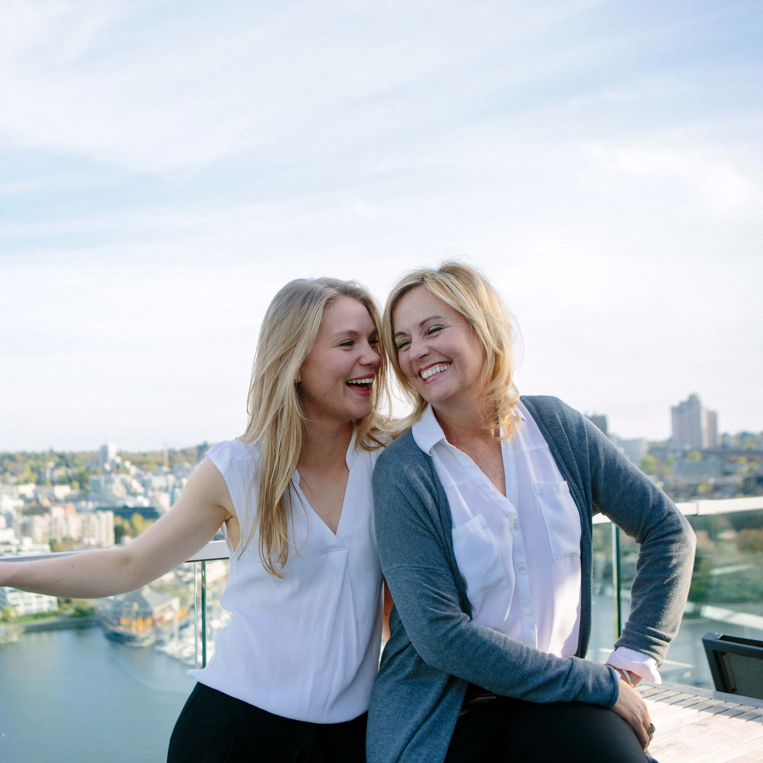 Two women are smiling and leaning close to each other on a balcony overlooking a city skyline; one woman has long blond hair and the other has shoulder-length blonde hair. The sky is partly cloudy.