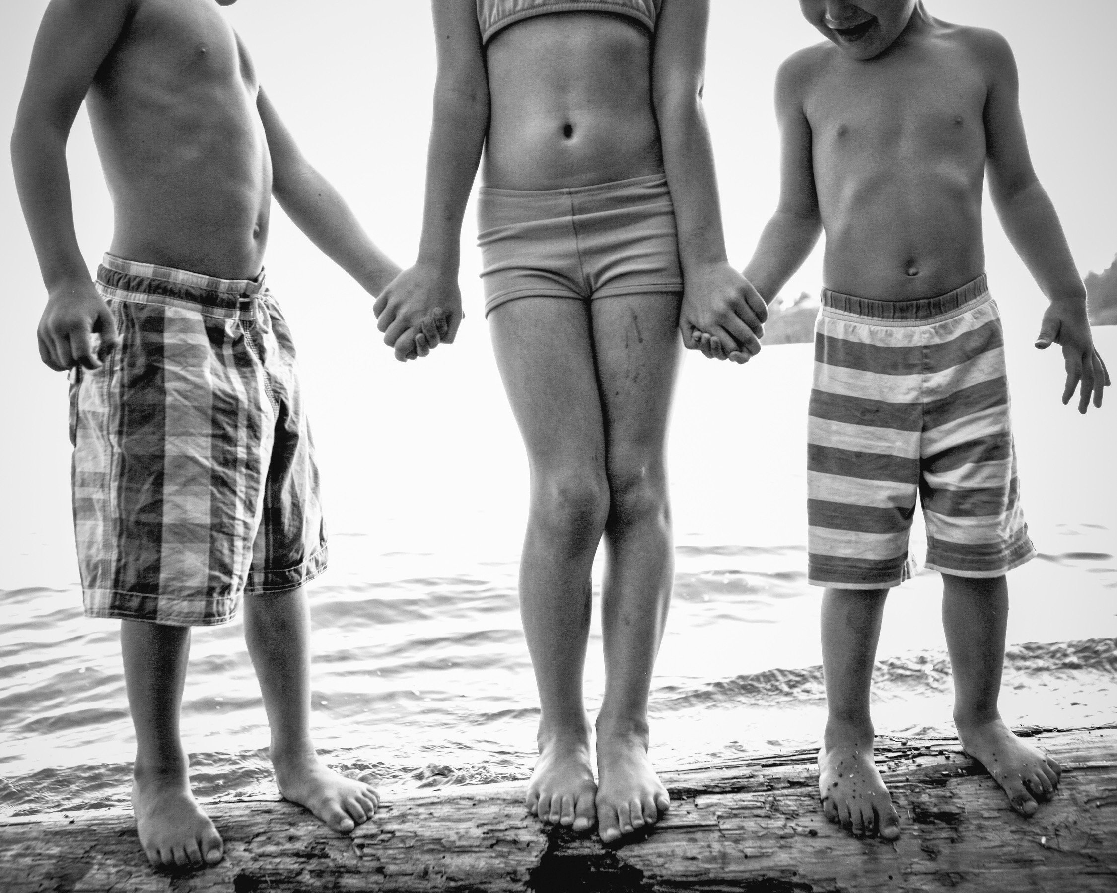 Three children standing arm in arm on the beach, holding hands, with water in the background. The photo is in black and white.