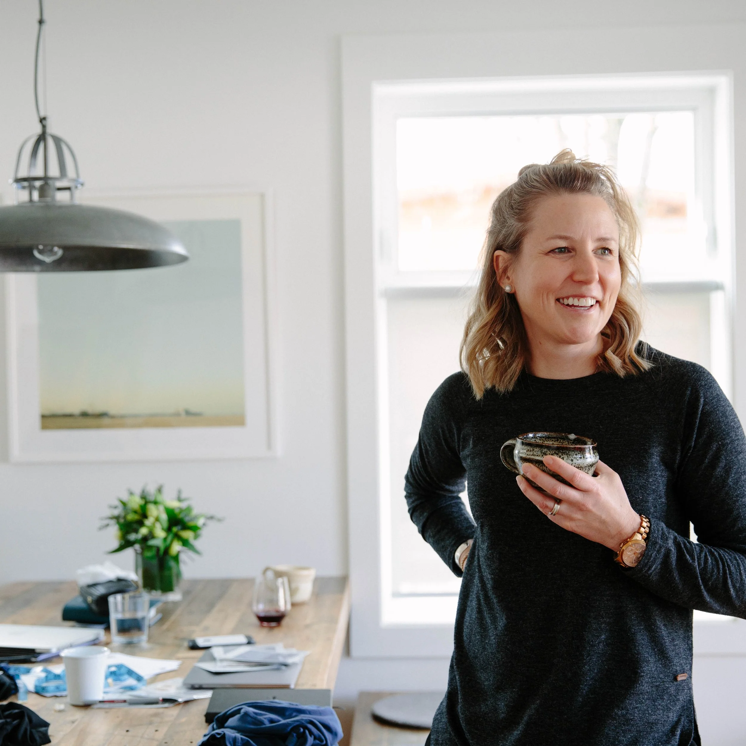 A smiling woman with light brown hair tied back, wearing a black top, holding a ceramic mug in her right hand, standing in a bright kitchen with a wooden table, plants, and various items on the table.