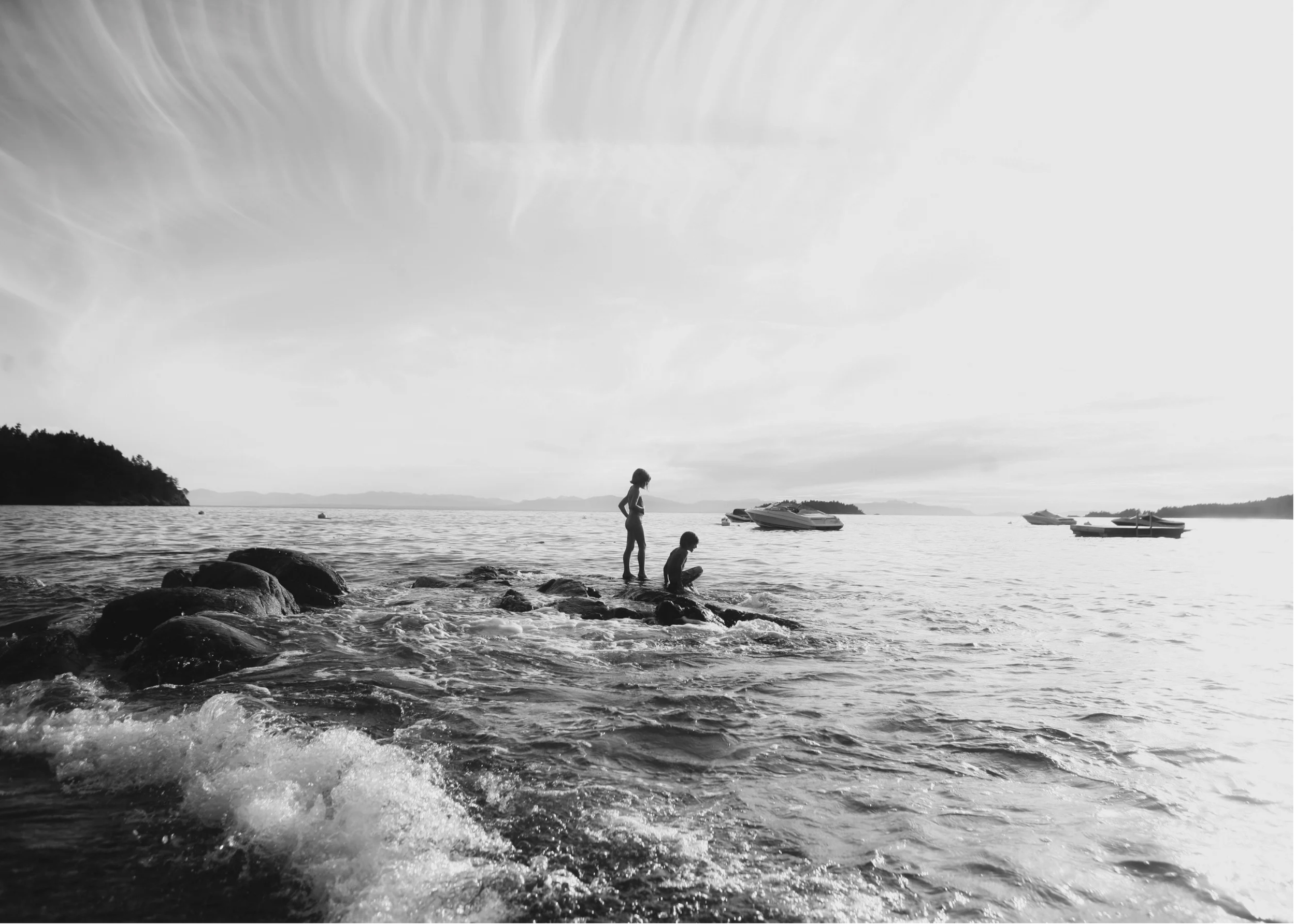A black and white photograph of a lakeside scene with two children, one standing and one sitting on rocks, near the water's edge. There are boats anchored in the water and a distant shoreline with trees. The sky has wispy clouds.