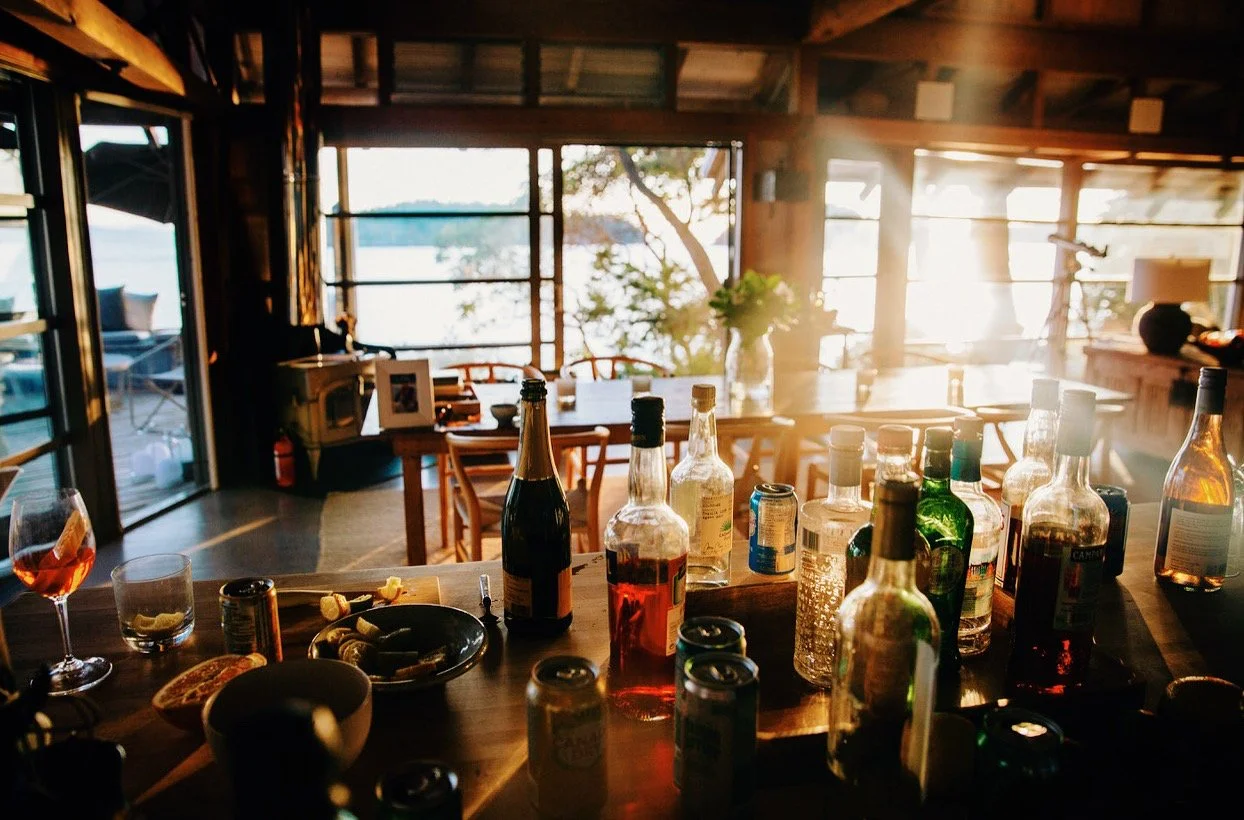 Interior of a rustic home with a wooden bar counter holding various bottles and drinks, large open windows letting in sunlight, and a dining area with chairs and a table in the background.