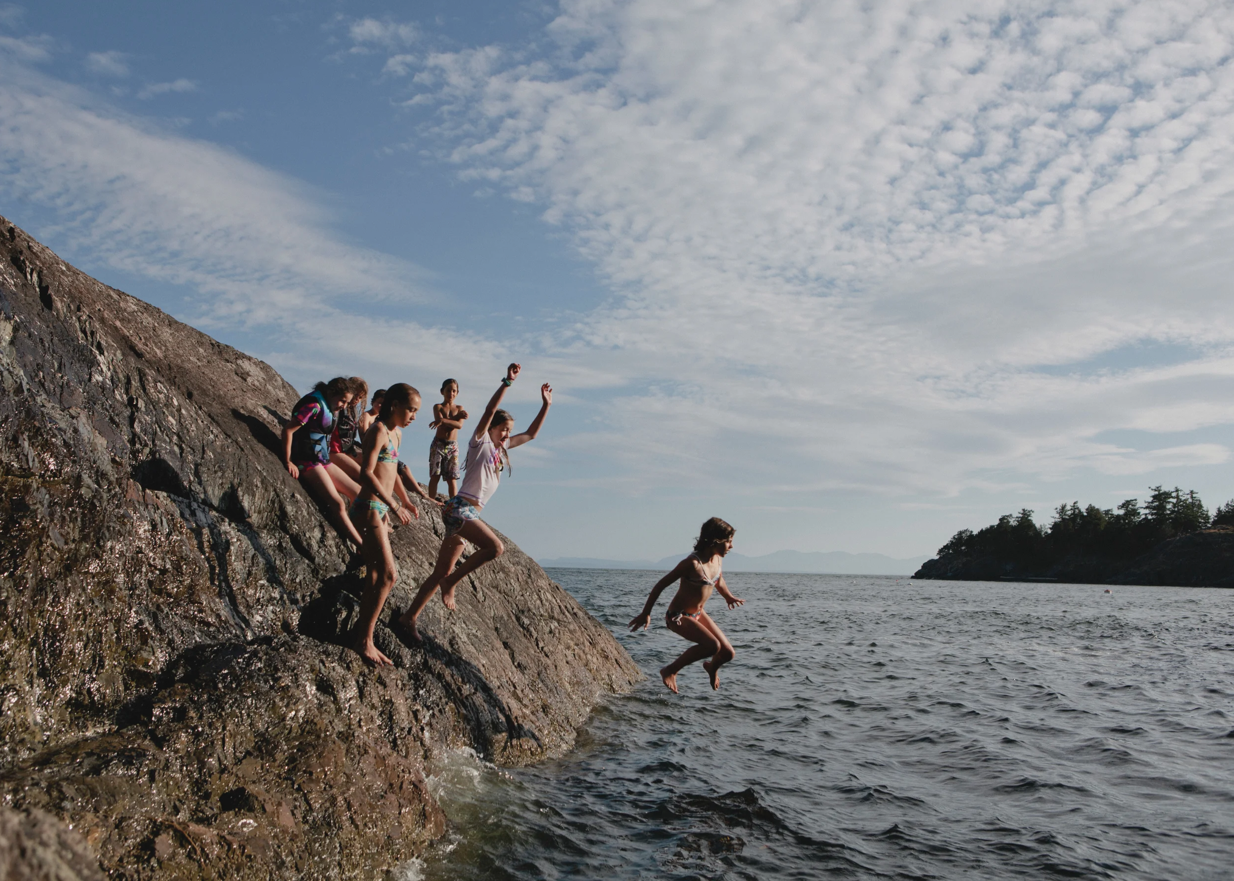 Children jumping off a rocky ledge into the water during daytime, with the sky partly cloudy and trees visible in the background.