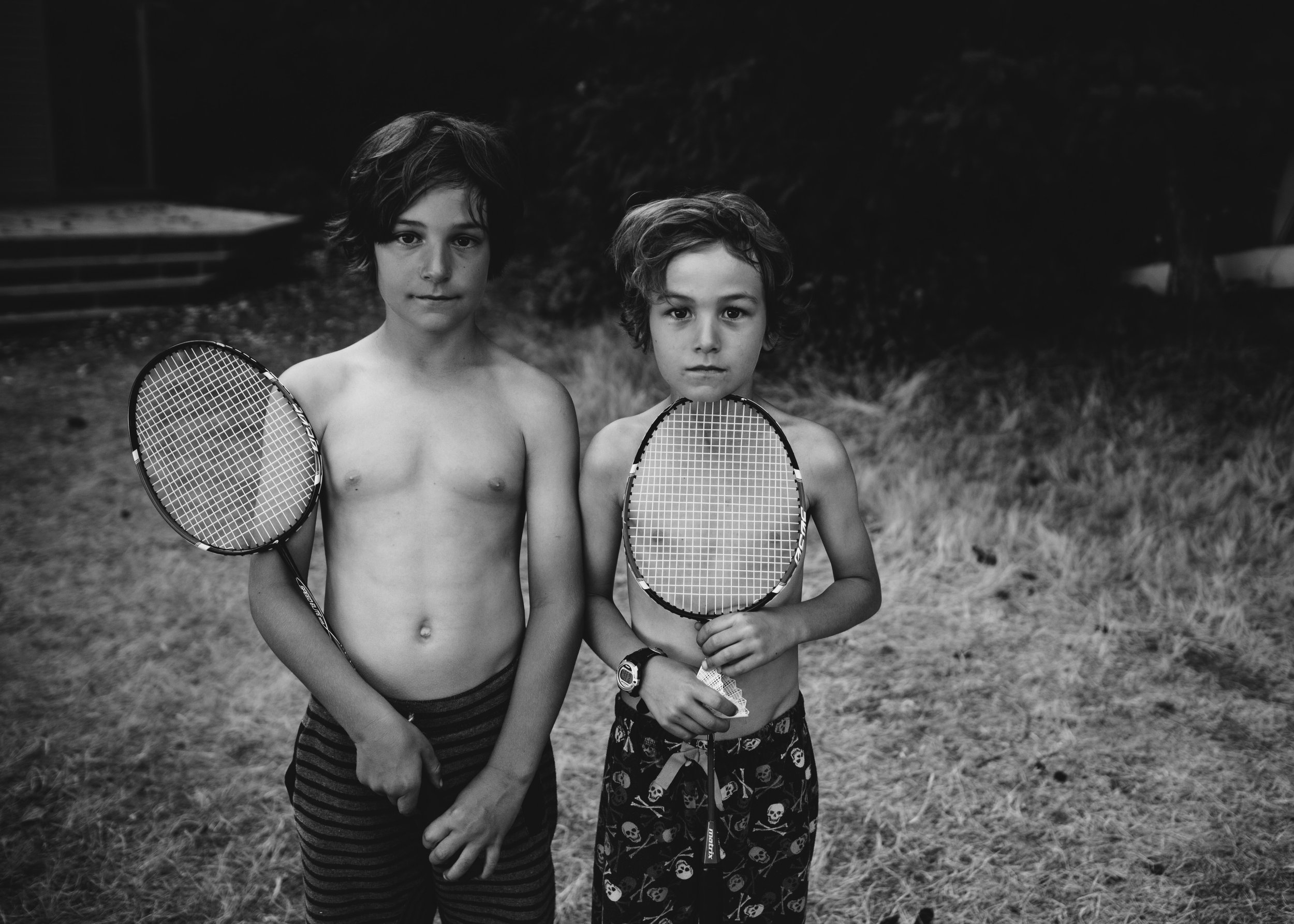 Two shirtless boys standing outdoors on grass, holding tennis rackets, looking directly at the camera. One has dark, wavy hair, striped shorts, and a watch, while the other has curly hair, skull-patterned shorts, and is wearing a wristwatch.