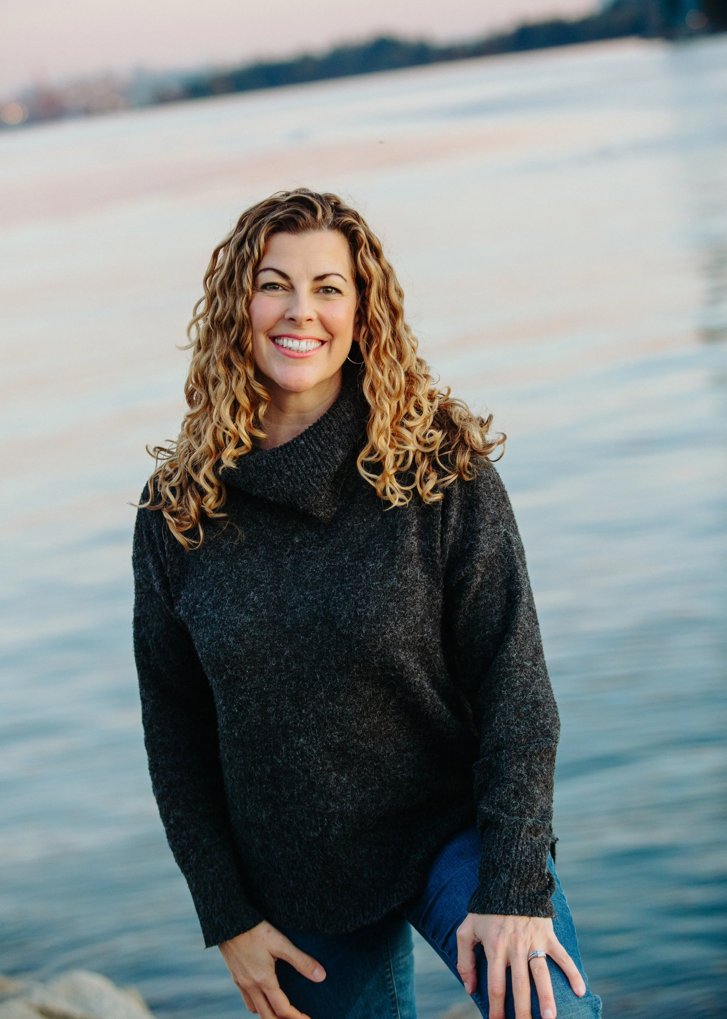 A woman with curly blonde hair smiling by a body of water during sunset, wearing a dark sweater and jeans.