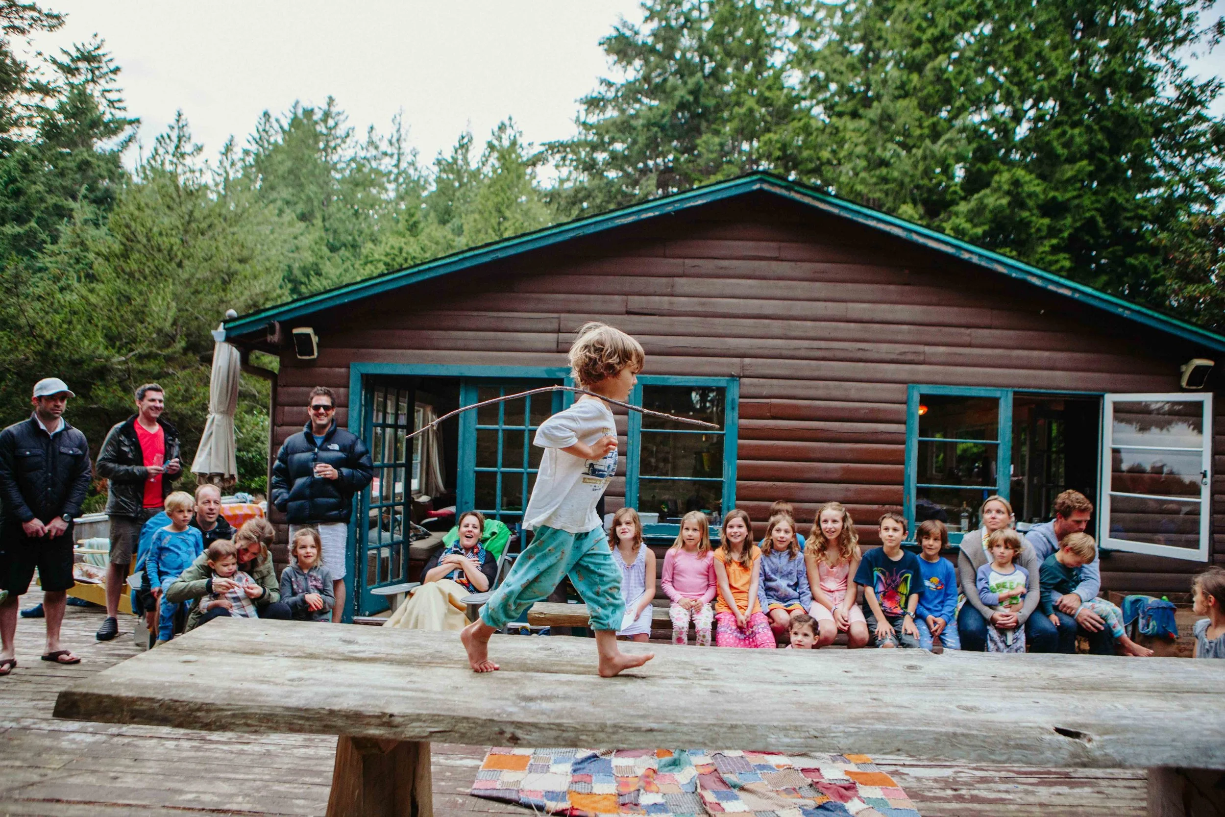 Children watching a performer in front of a crowd outside near a wooden house surrounded by trees.