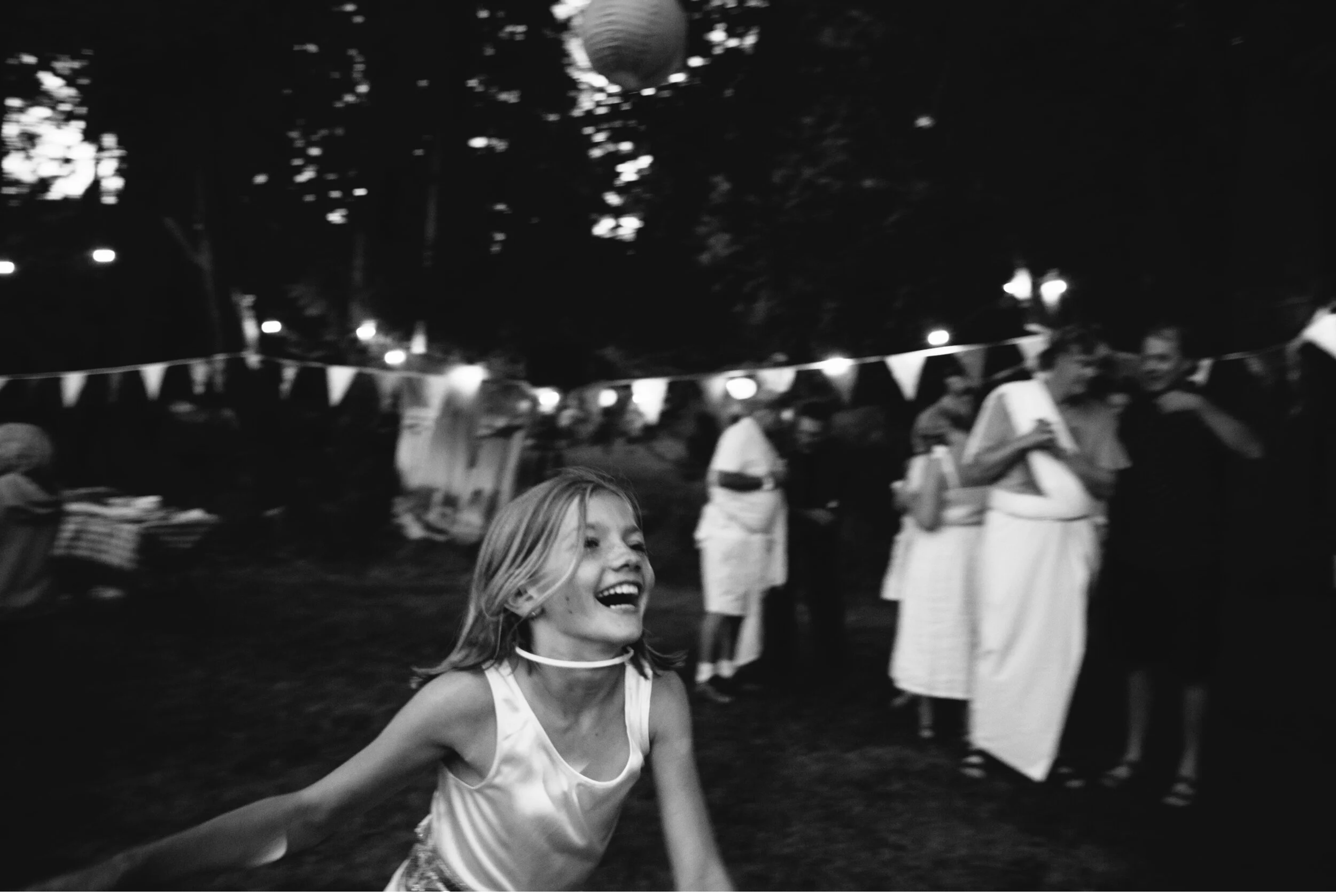 A young girl with blonde hair smiling and running at an outdoor event during the evening. In the background, there are people walking and standing, some holding papers, and decorative bunting hanging overhead.