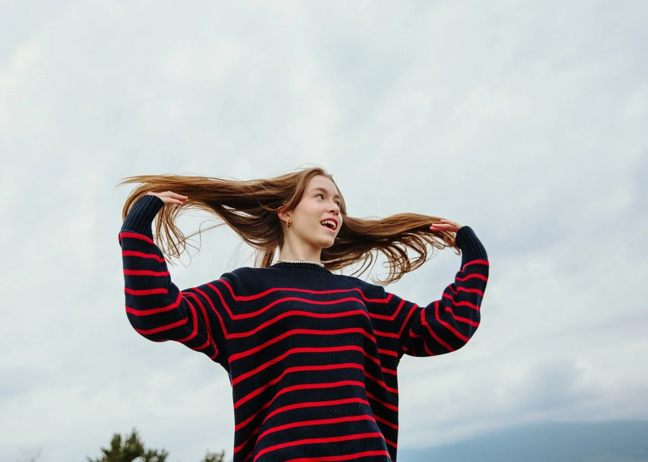 Young woman with long hair in a striped sweater, standing outdoors against a cloudy sky, with her hair flowing in the wind.