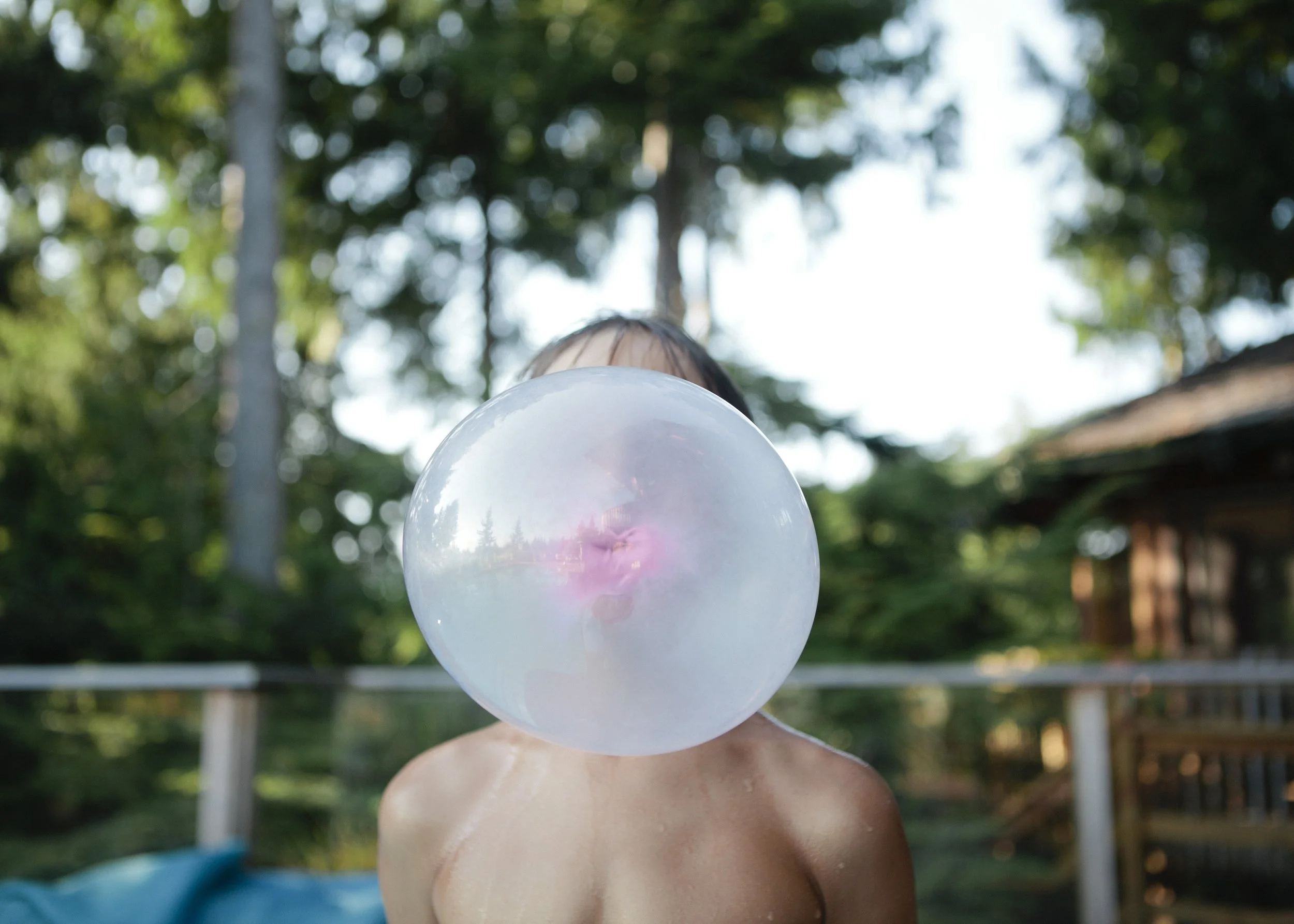 Child blowing a bubble with gum outdoors, with trees and a wooden structure in the background.