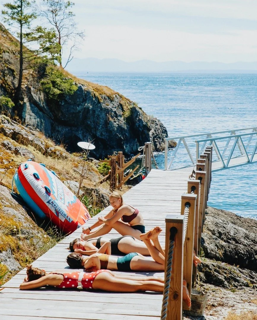 Three women relaxing on a wooden dock by the water, with an inflatable boat nearby and rocky cliffs in the background, during daytime.