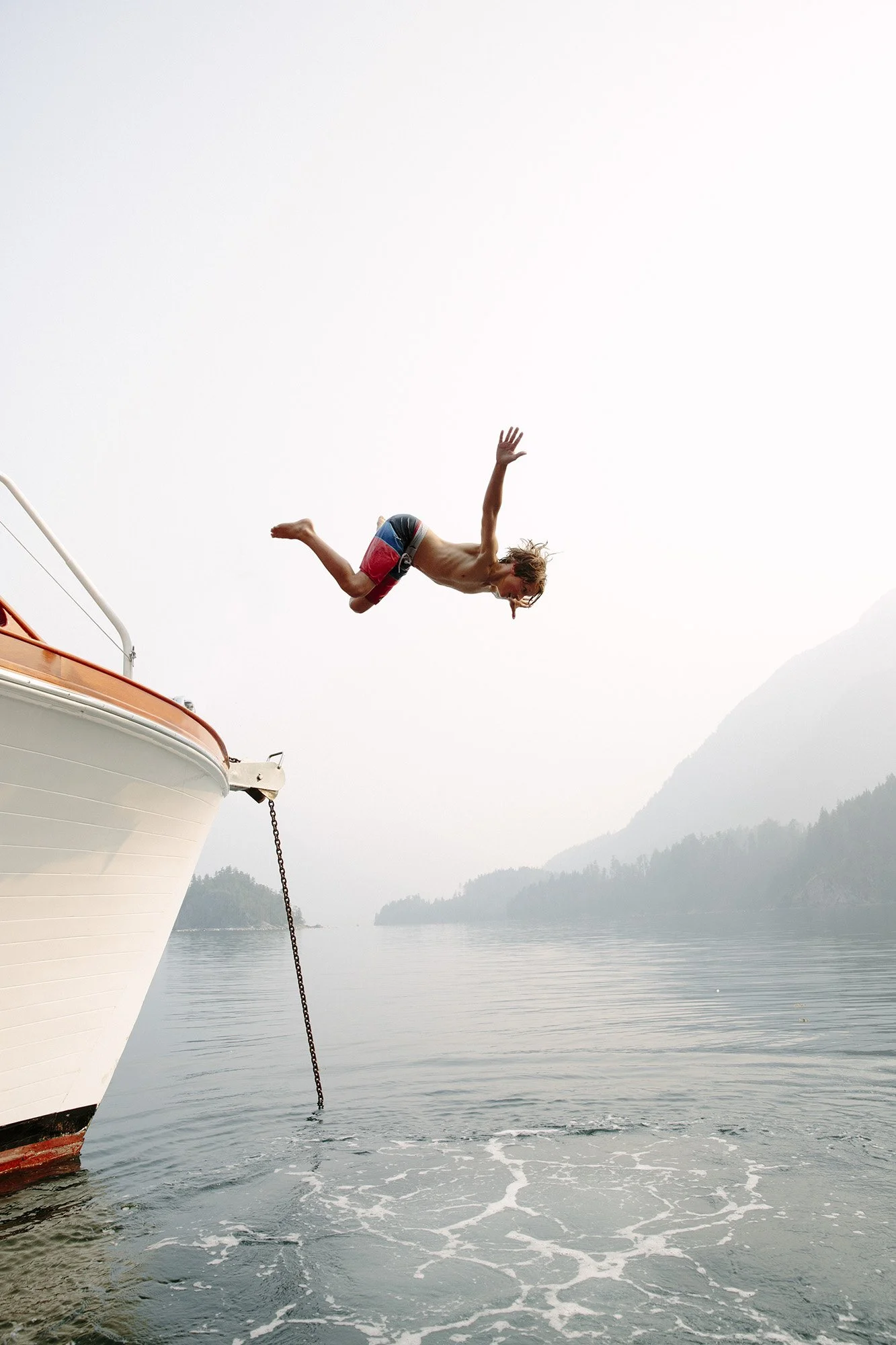 A person jumping off the bow of a boat into the water, surrounded by foggy mountains and calm water.