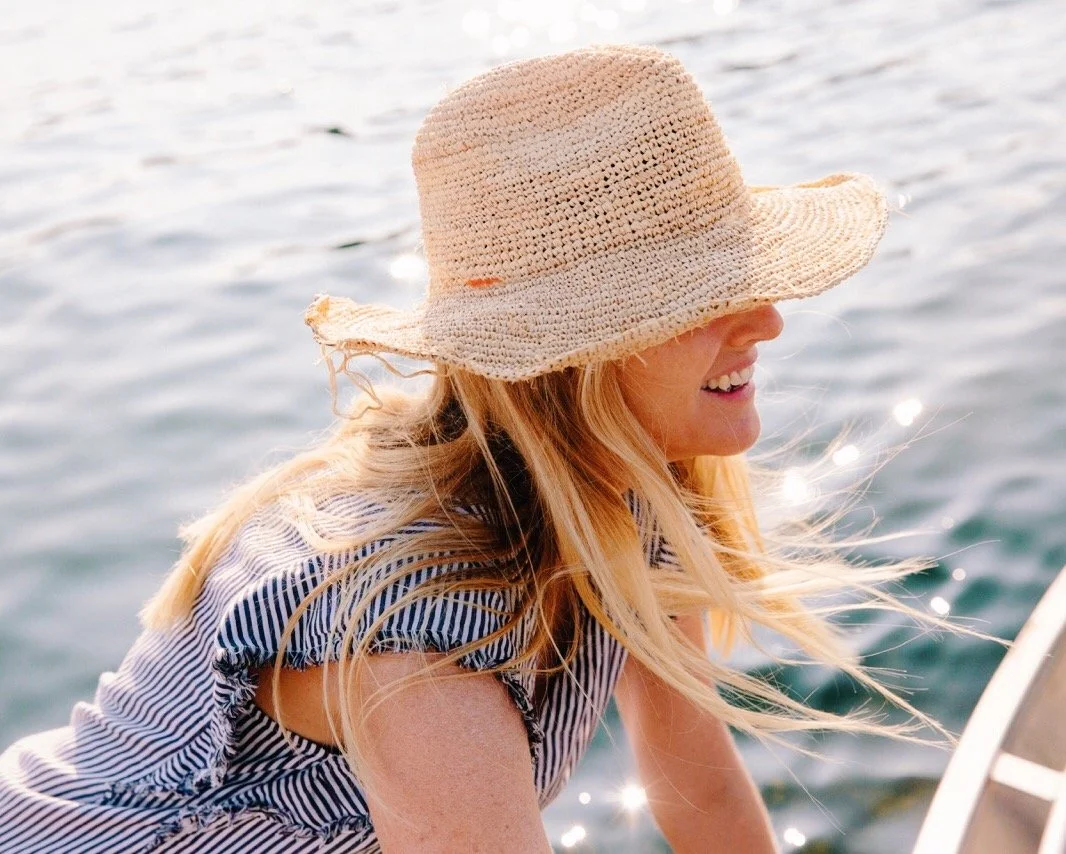 A woman with long blonde hair wearing a straw hat and a striped sleeveless top, smiling by the water.