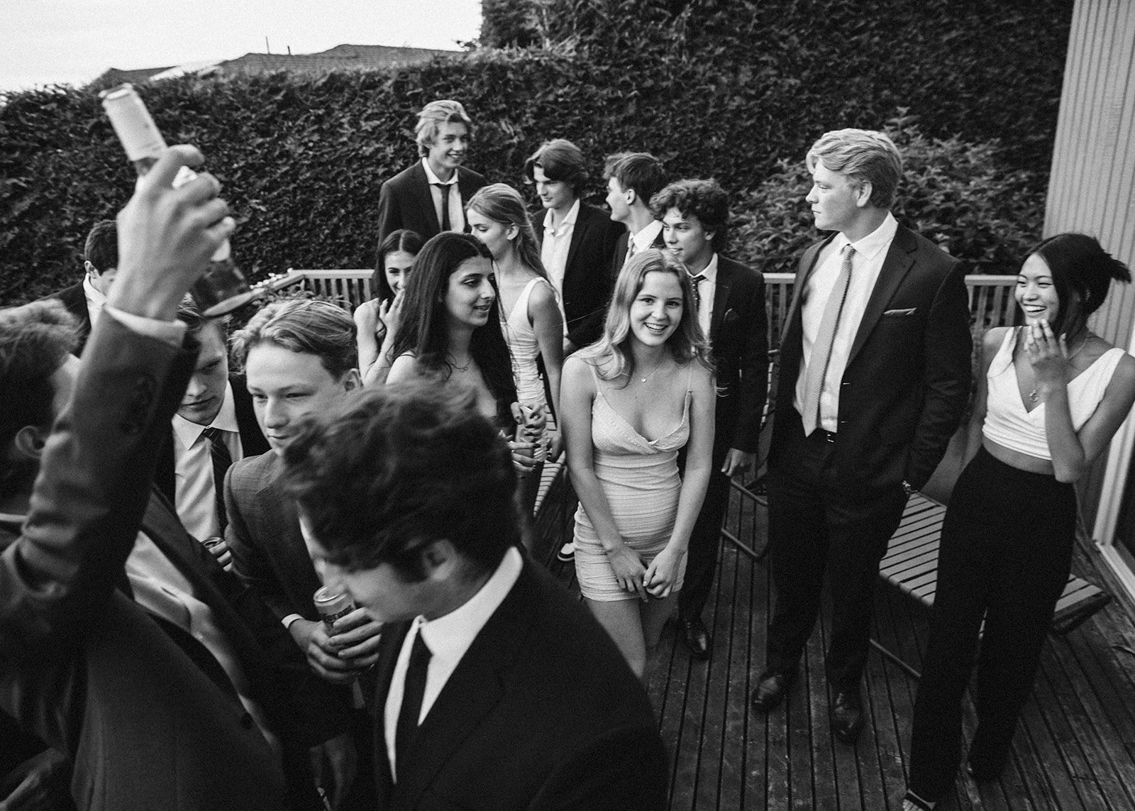 Group of young adults at a social gathering on a deck, some smiling and chatting, dressed in semi-formal attire, with a background of trees and a building.