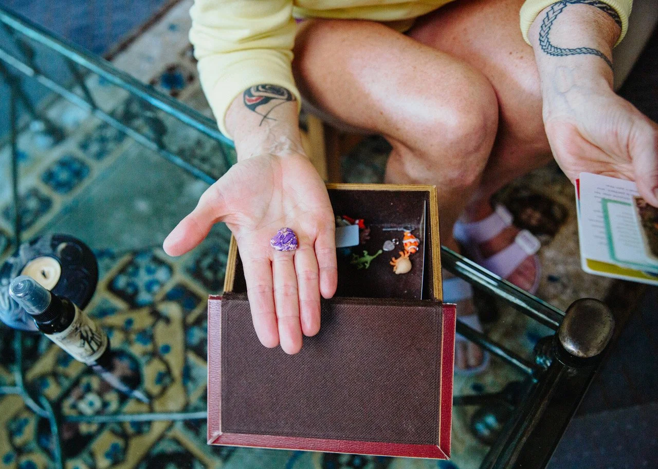 A person with tattoos on their arms is showing a purple ring on their finger while holding a small box with colorful figurines or toys inside. The person is sitting at a glass table.