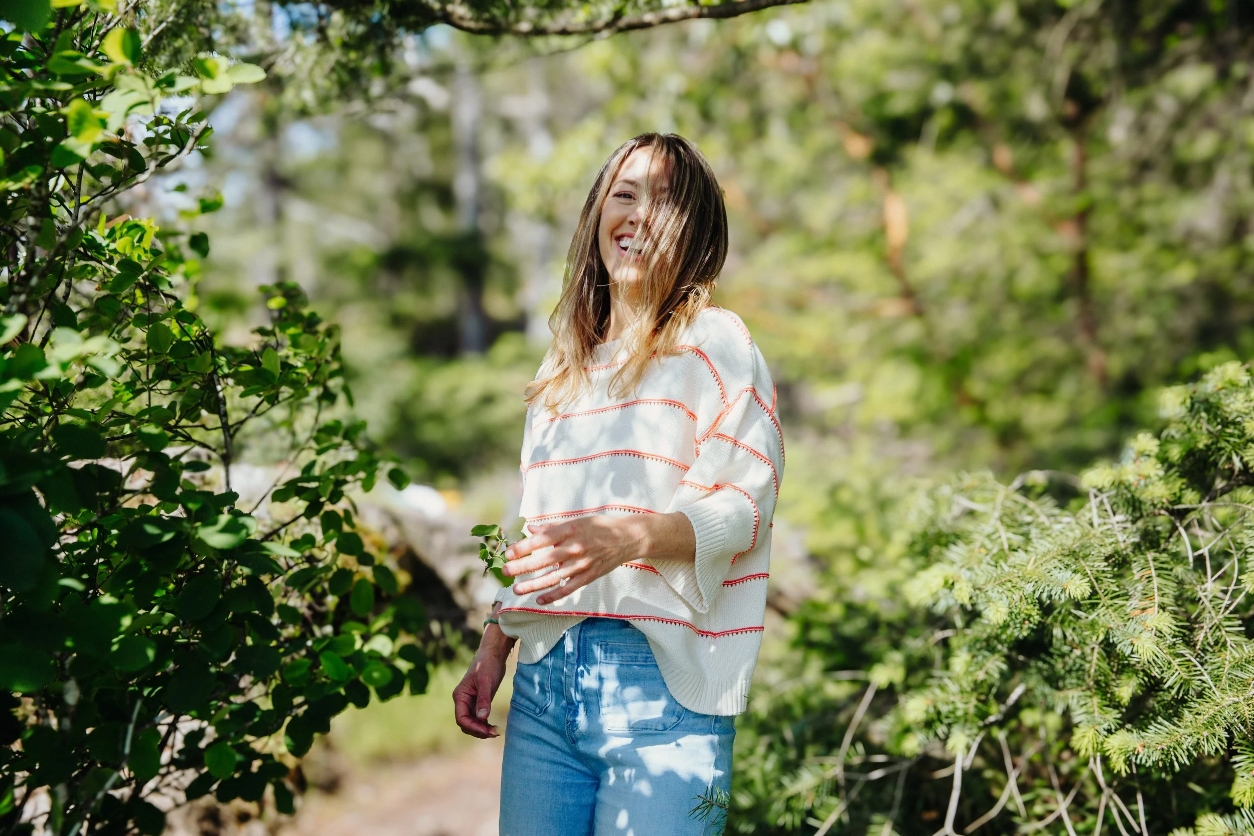 A woman with long hair, smiling and looking at the camera, stands outdoors in a lush green garden or park with sunlight filtering through trees. She is wearing a white sweater with red stripes and blue jeans.