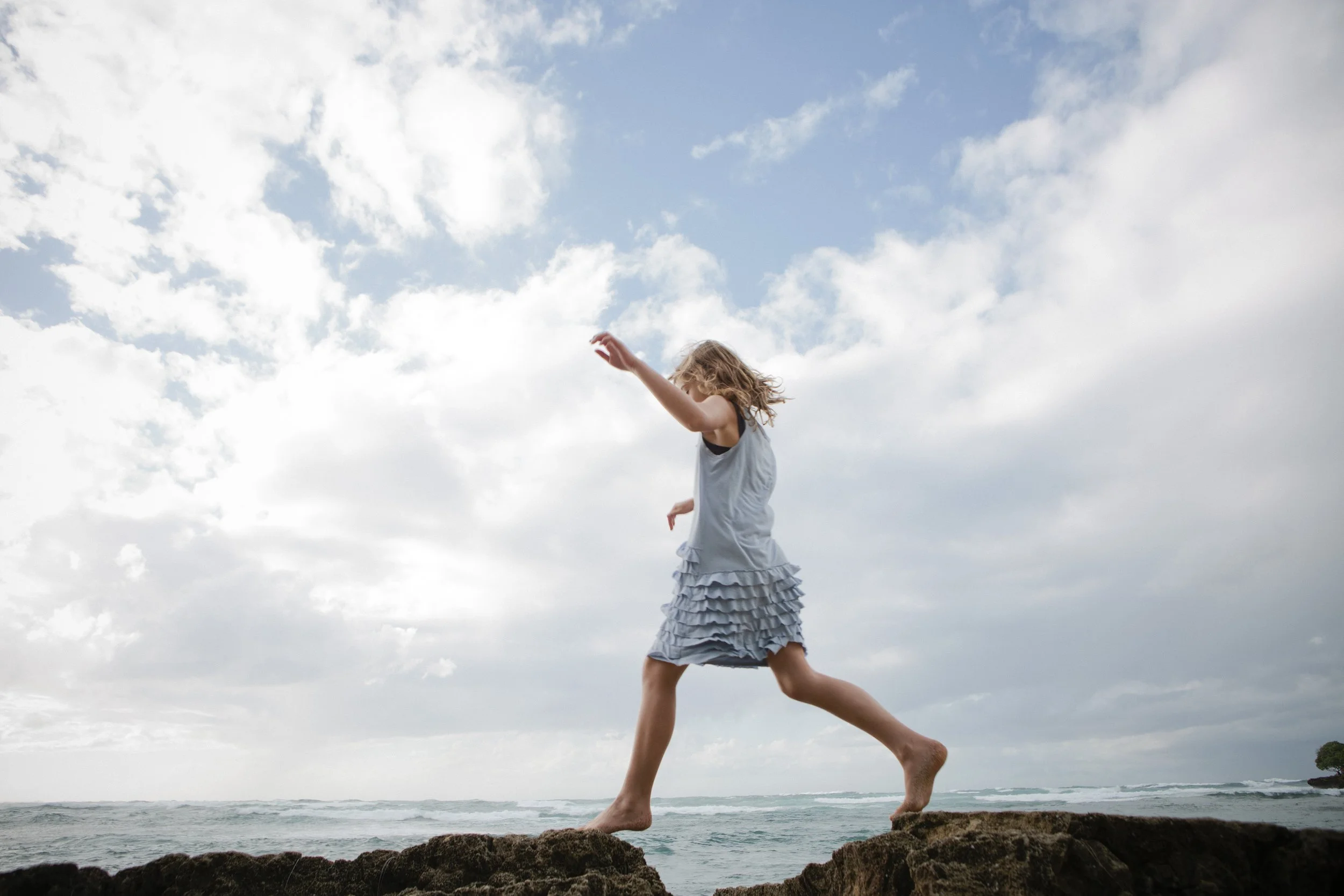 A girl in a light blue ruffled dress balancing barefoot on rocks by the ocean, with a partly cloudy sky overhead.