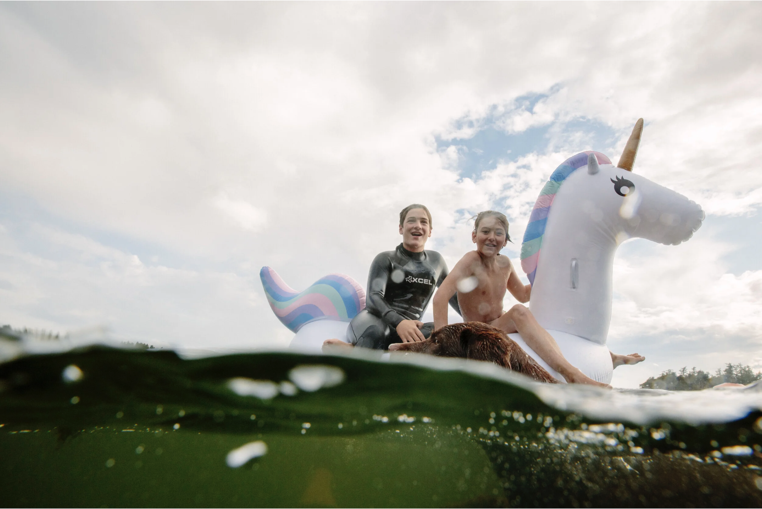 Two children and a dog riding on an inflatable unicorn float in a lake, viewed from underwater with a cloudy sky above.