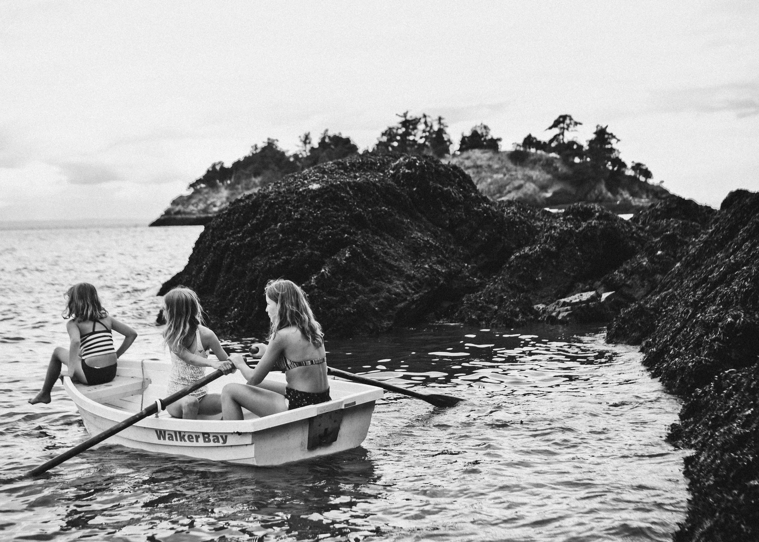 Four girls in swimsuits rowing a small boat labeled 'Walker Bay' on a body of water, with a rocky island and trees in the background.