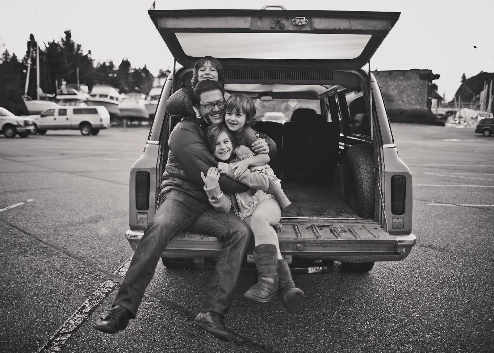 A man and four children sit in the open trunk of a car, all smiling and hugging each other, in a parking lot with boats and trees in the background.
