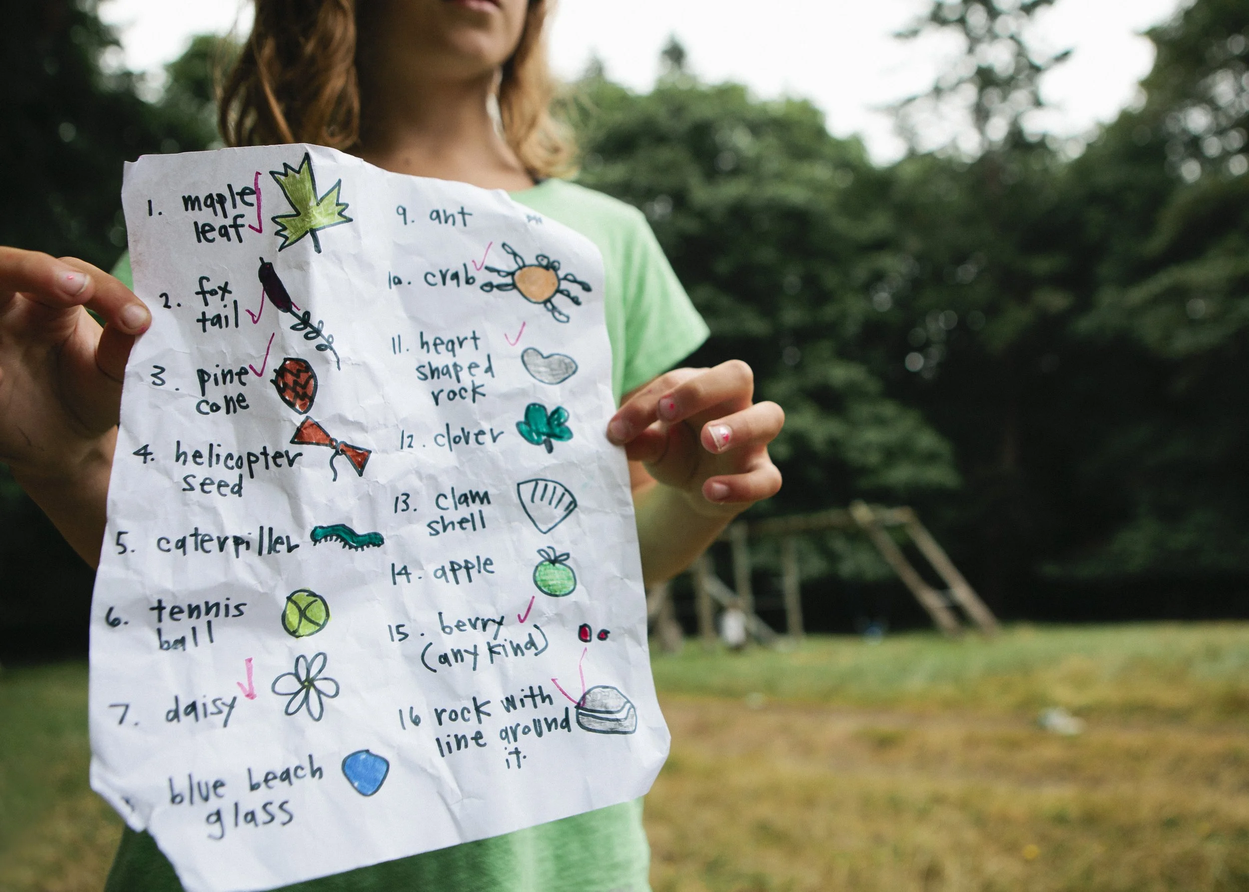 A person holding a handwritten list with colorful drawings, outdoors with trees in the background.