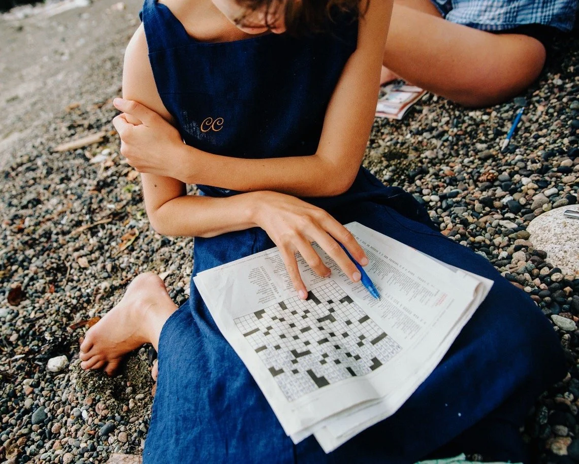 Person sitting on rocky ground working on a crossword puzzle with a blue pen.
